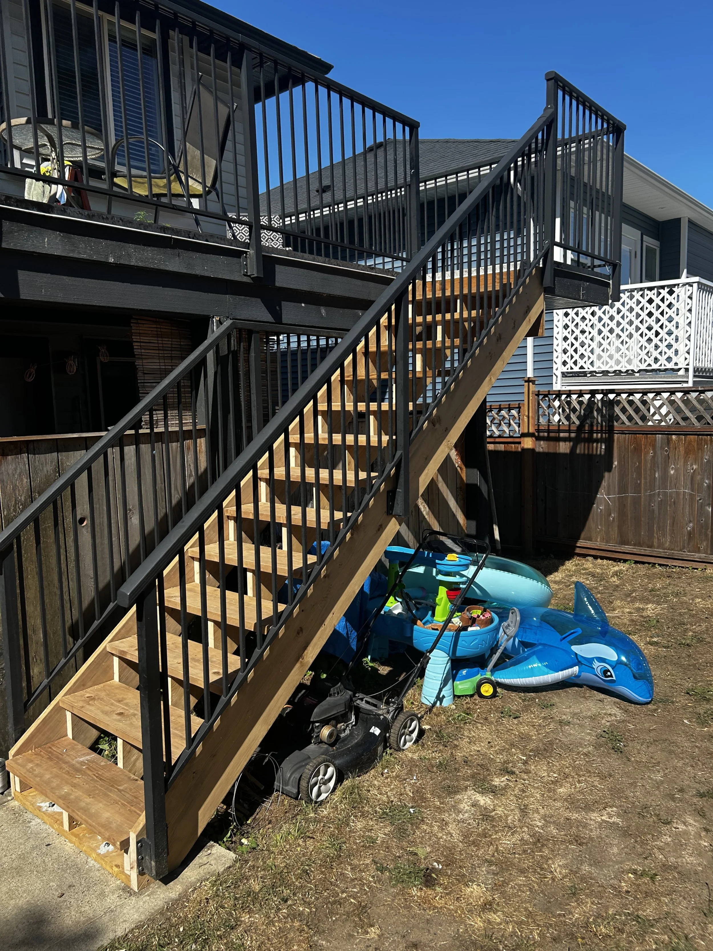 Backyard with newly installed wooden staircase and black metal railing, leading to an upper deck with outdoor furniture, children's toys including a blue dolphin pool float, a toy lawn mower, and a sandbox, on dry grass and dirt ground beside a wooden fence.