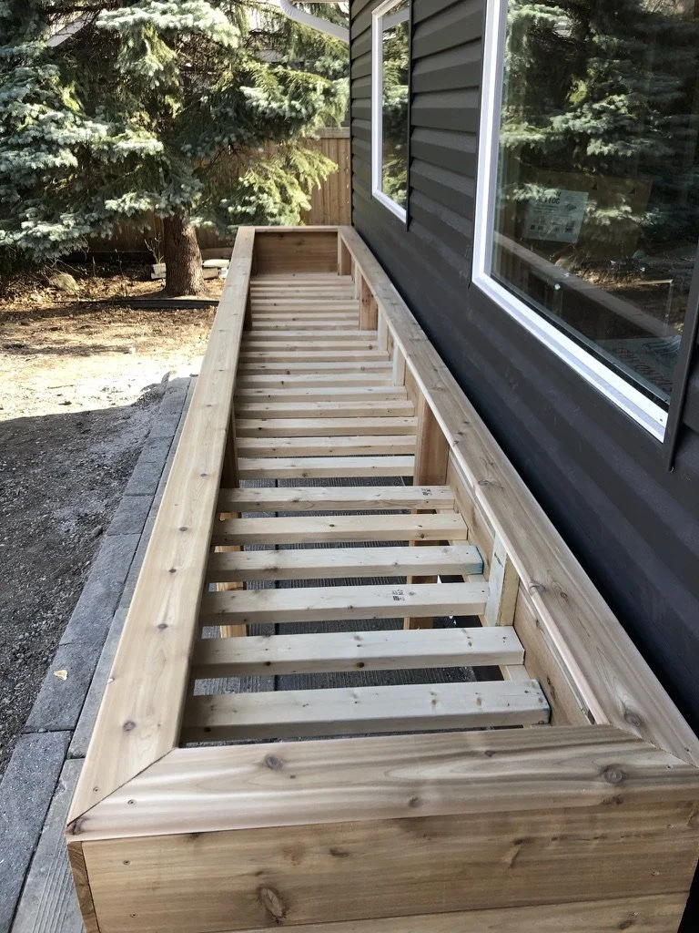 Wooden deck framing under construction next to a house with dark siding and large windows, with trees in the background.
