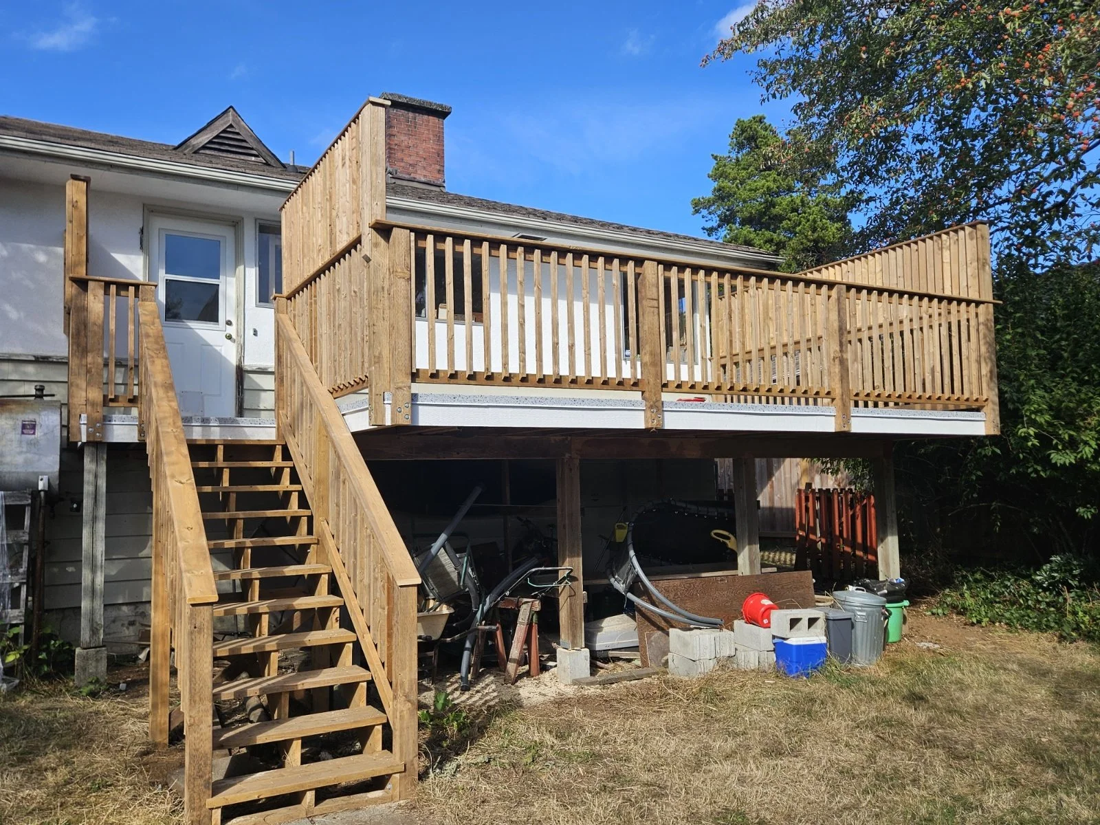 Construction site of a new wooden deck attached to the back of a house, with stairs leading up to it and a railing around the perimeter, under a clear blue sky.