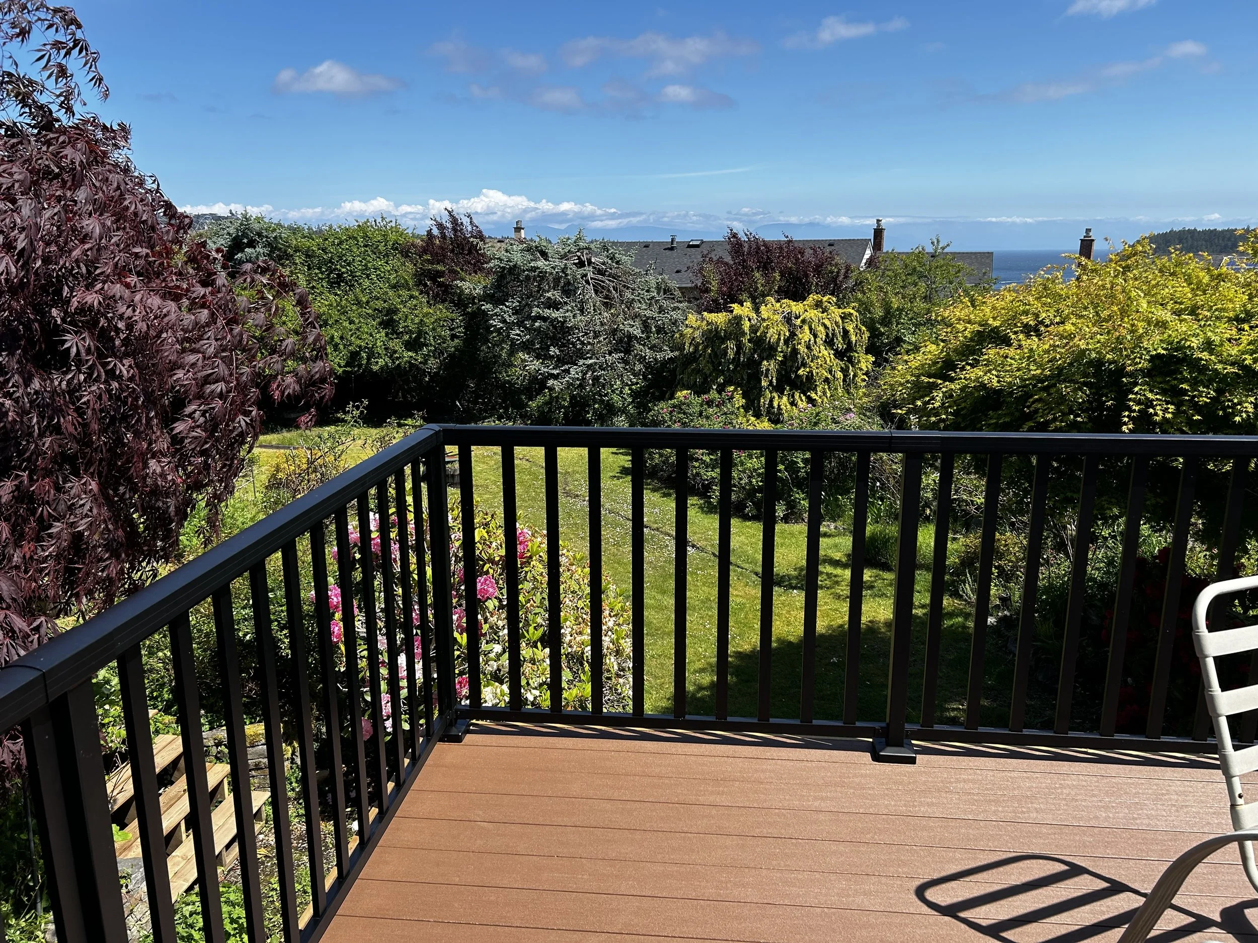 View of a backyard garden with green grass, colorful trees, shrubs, and flowers, seen from a wooden deck with black railing on a sunny day with a blue sky and some clouds.