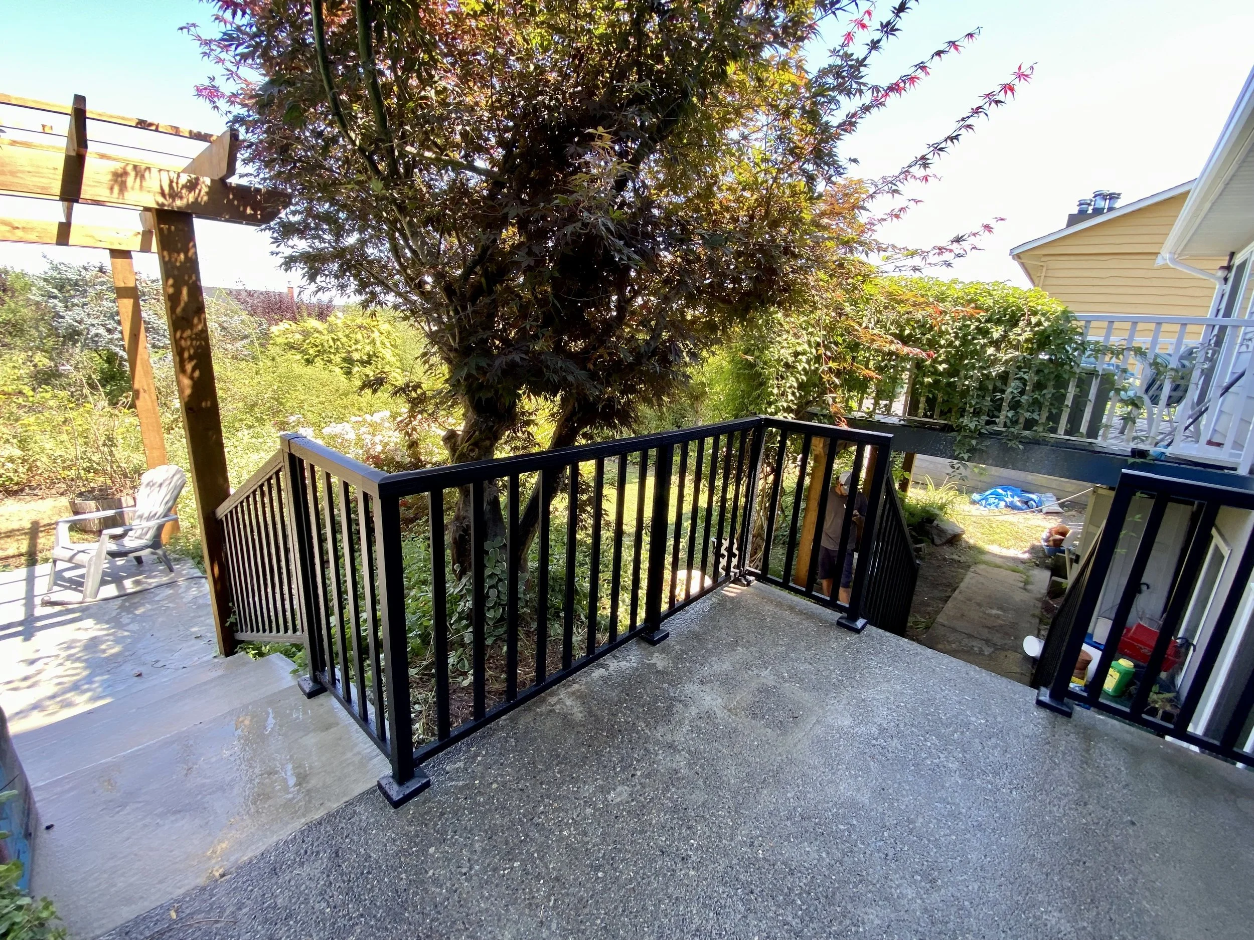 View of a backyard patio with concrete flooring, black metal railing, trees, and neighboring houses under a sunny sky.