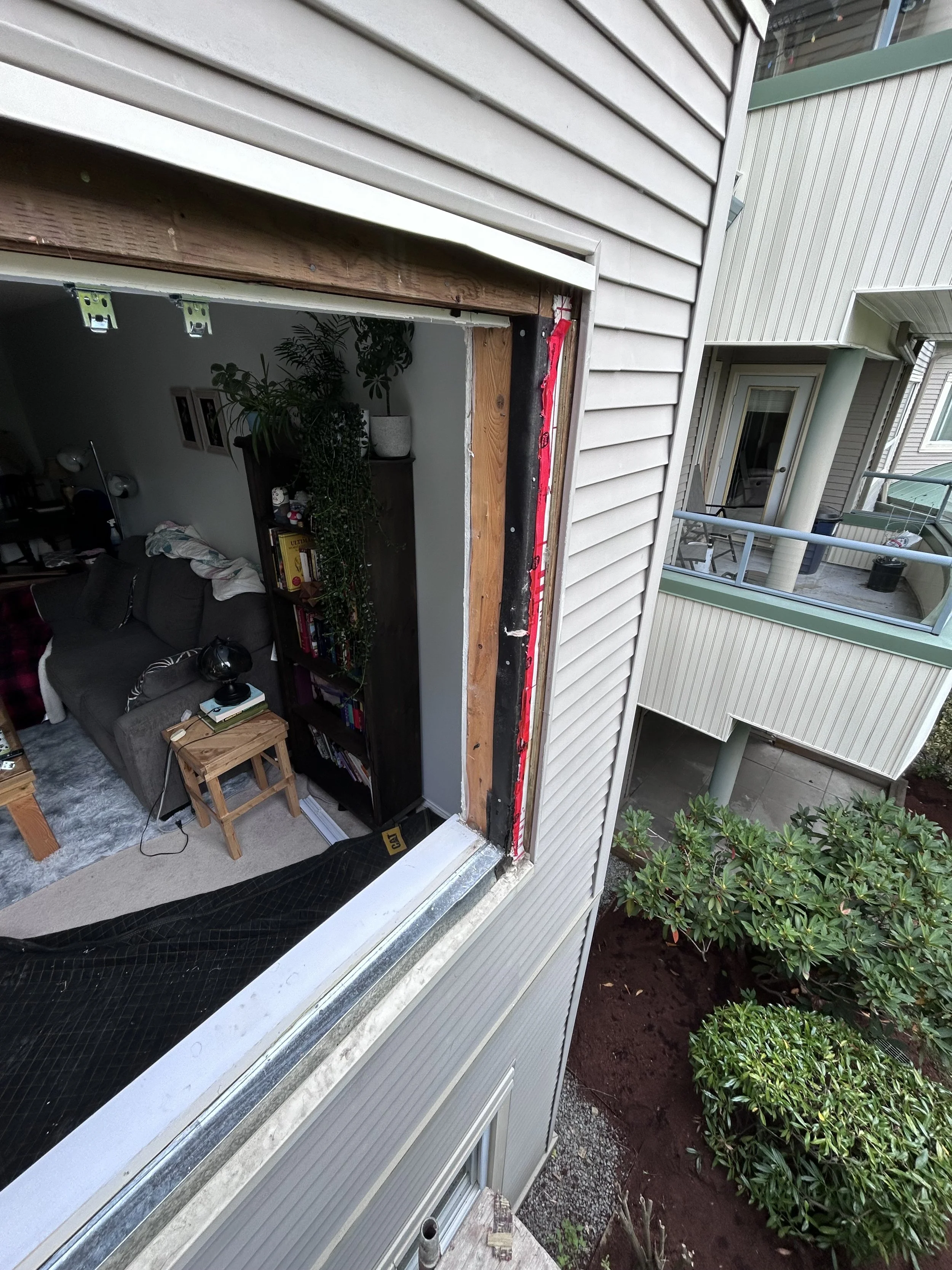 Photo of a building under renovation showing a partially removed window frame, with a view into a living room containing a gray sofa, a small wooden side table, a bookshelf, and indoor plants. The building's exterior siding is beige, and neighboring apartment units with balconies are visible.