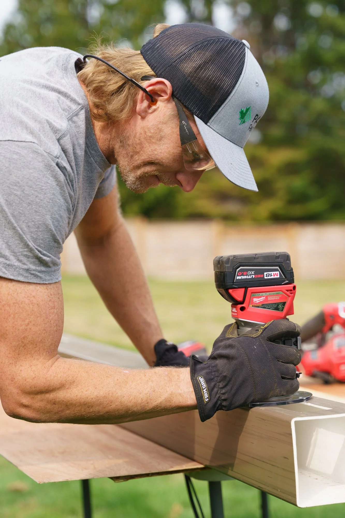 A man wearing safety glasses, a baseball cap, and black gloves is using a cordless power drill on a piece of wood outdoors during daytime.