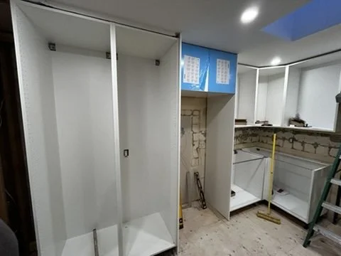 Kitchen area under renovation with open white cabinets, a partially built wall, and construction tools present.