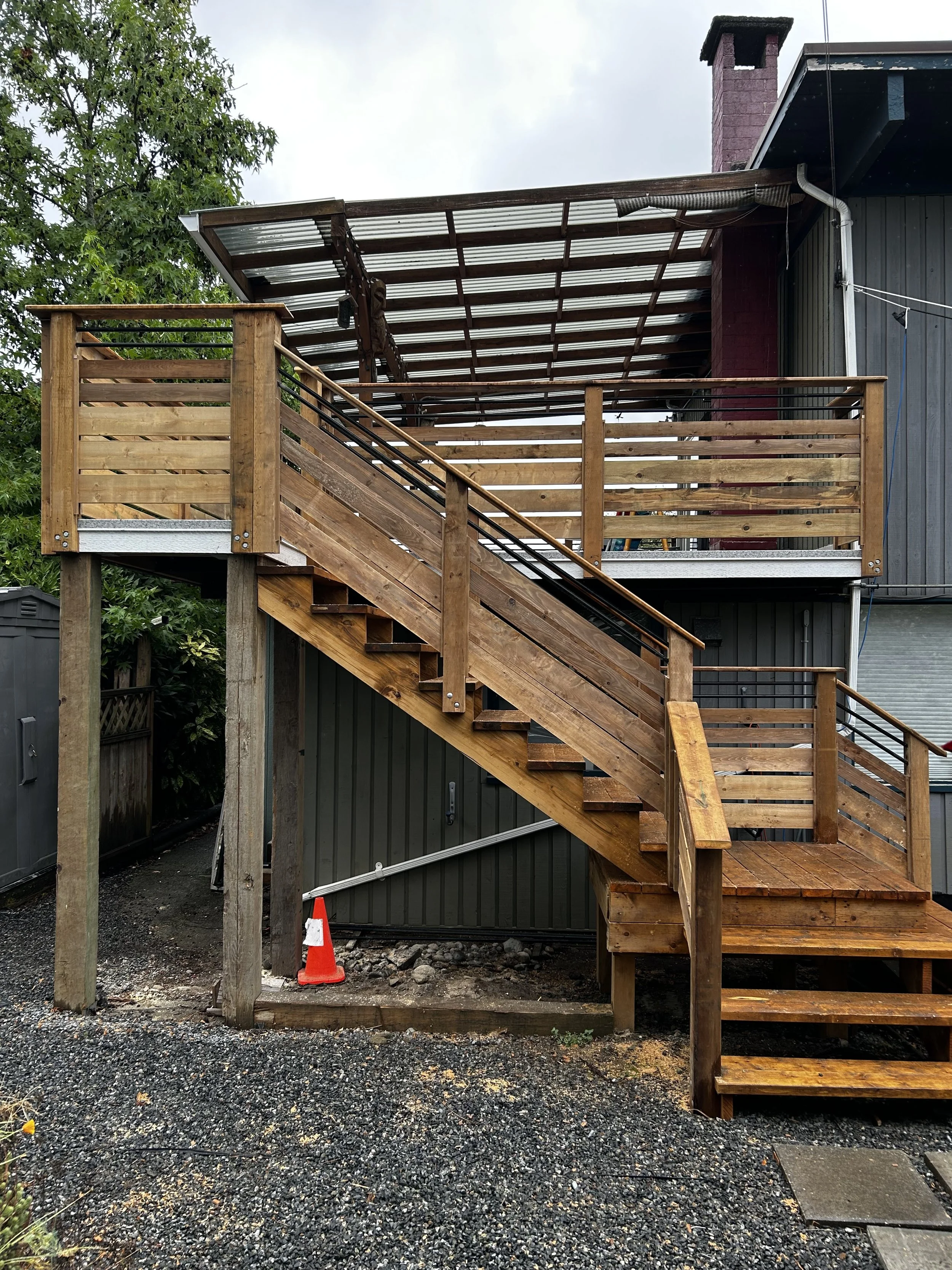 Newly built wooden outdoor staircase and balcony attached to a house with black metal railings, a metal roof, and a large red brick chimney.