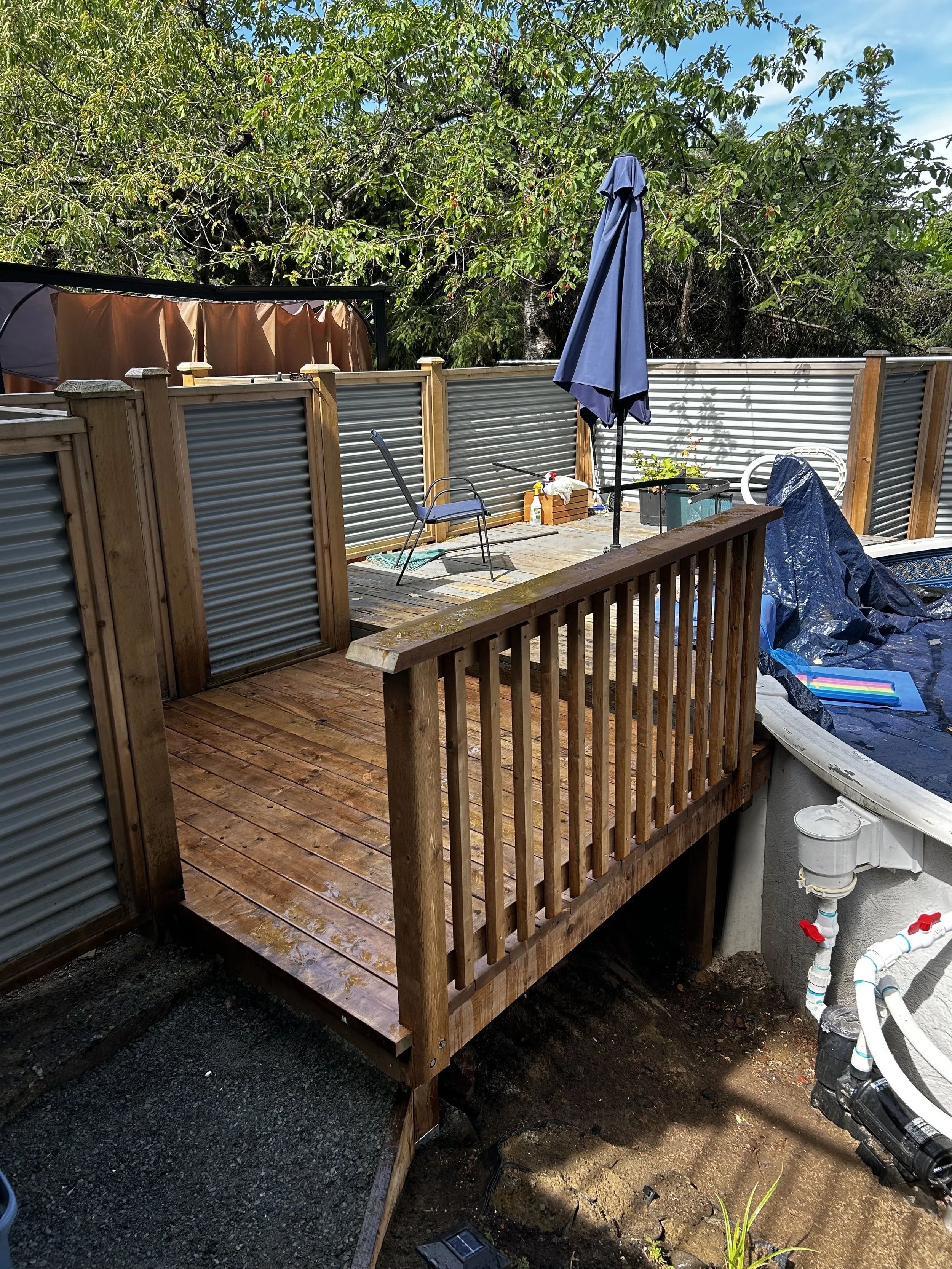 A wooden deck with a railing attached to a above-ground pool with a tarp cover. There is a closed blue umbrella, chairs, and gardening tools on the deck. A backdrop of trees and a metal fence surrounds the area.