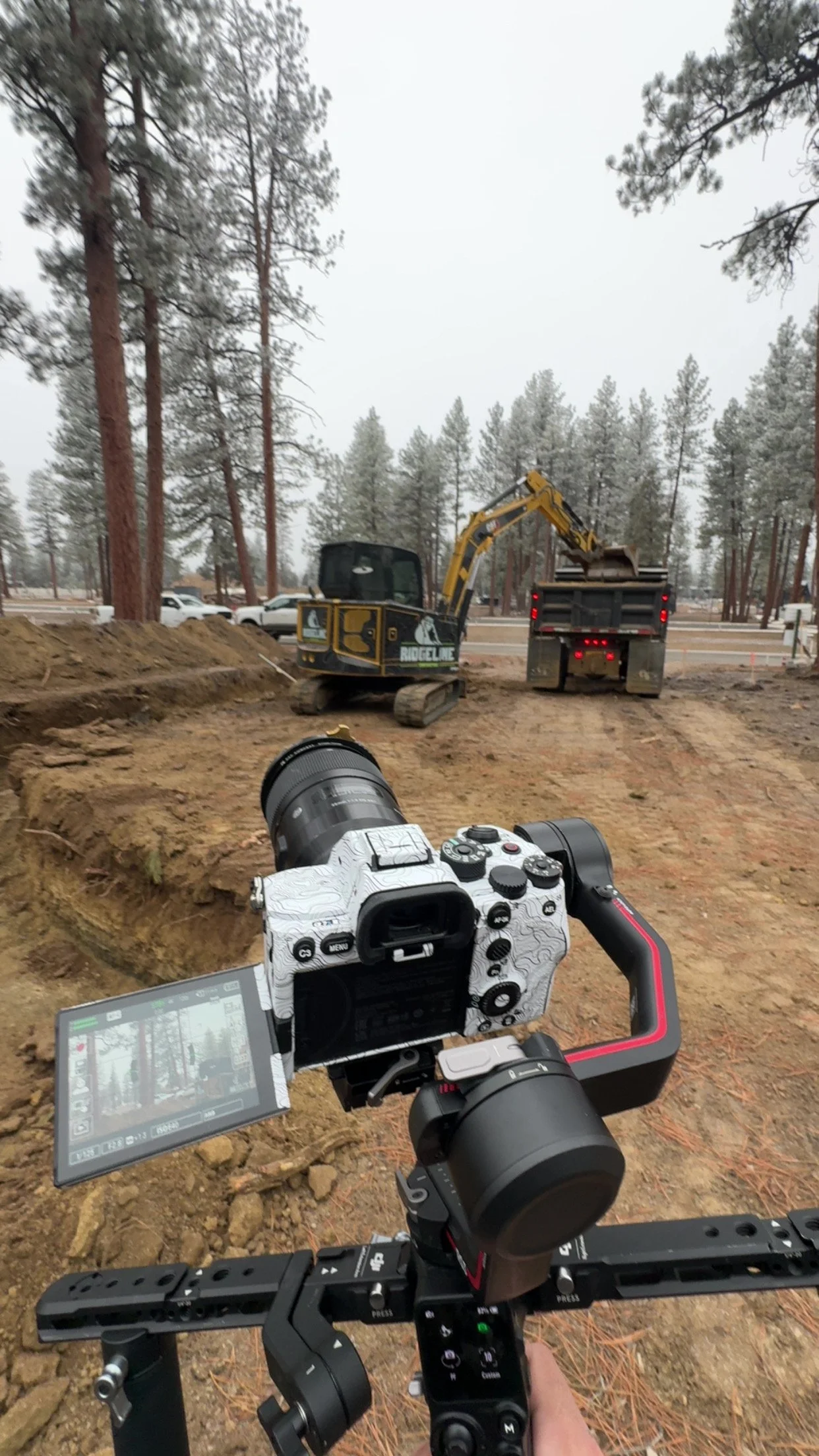 Construction scene with a camera in the foreground, a mini excavator, and a dump truck at a muddy construction site surrounded by tall trees, under an overcast sky.