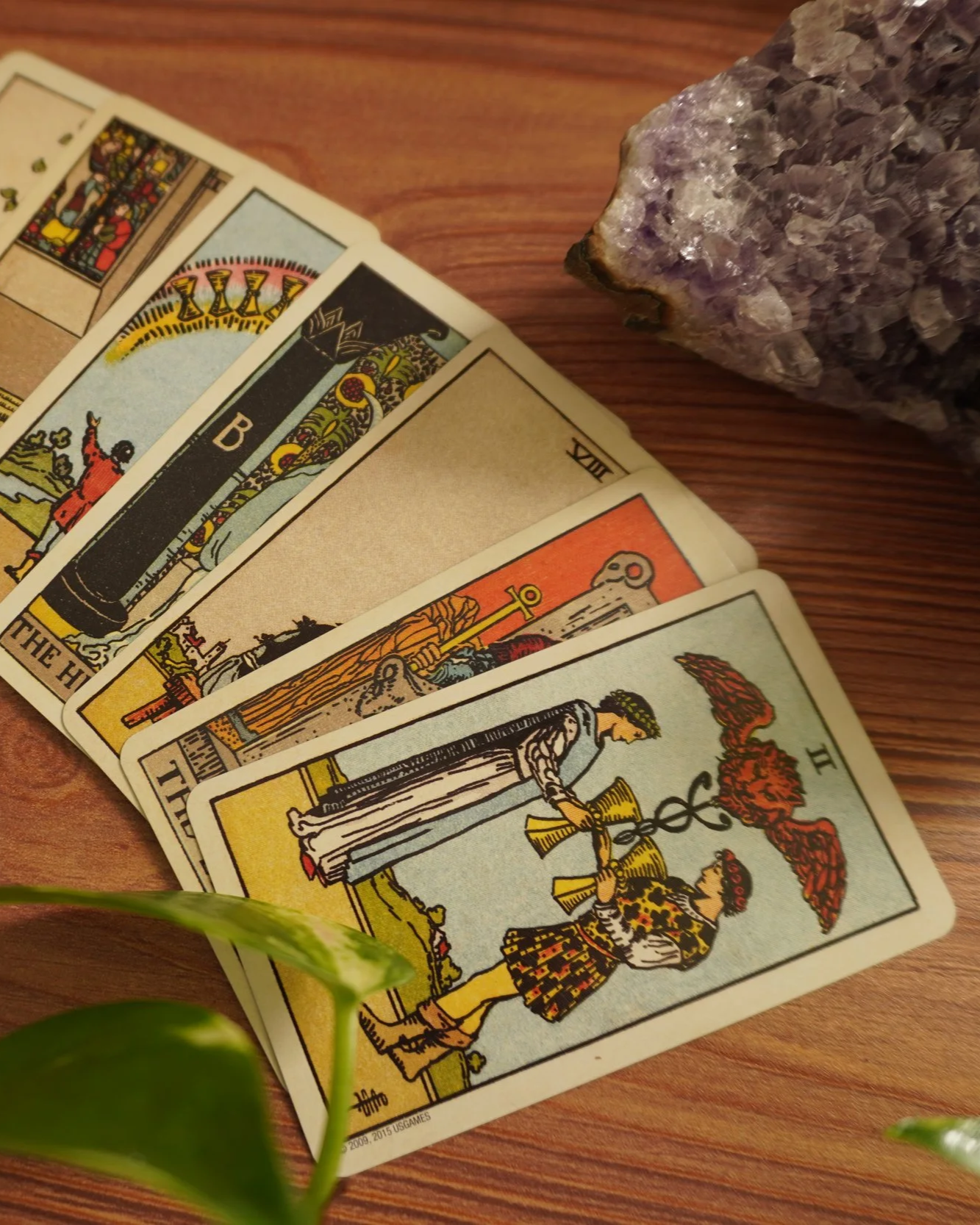 A deck of tarot cards spread out on a wooden surface next to a purple crystal and some green leaves in the foreground.