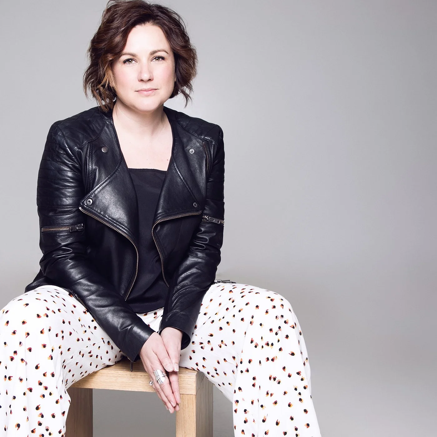 A woman sitting on a wooden stool, wearing a black leather jacket, a black top, and white pants with a colorful polka dot pattern, posing against a plain grey background.  She looks like a leader who would help develop business content and strategy.