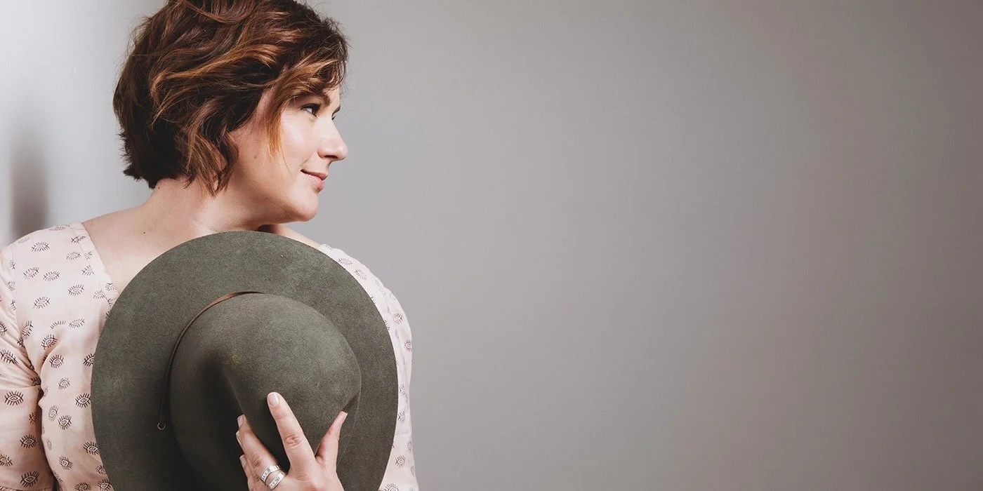 A woman with short reddish-brown hair holding a gray hat, looking to her right against a plain wall.