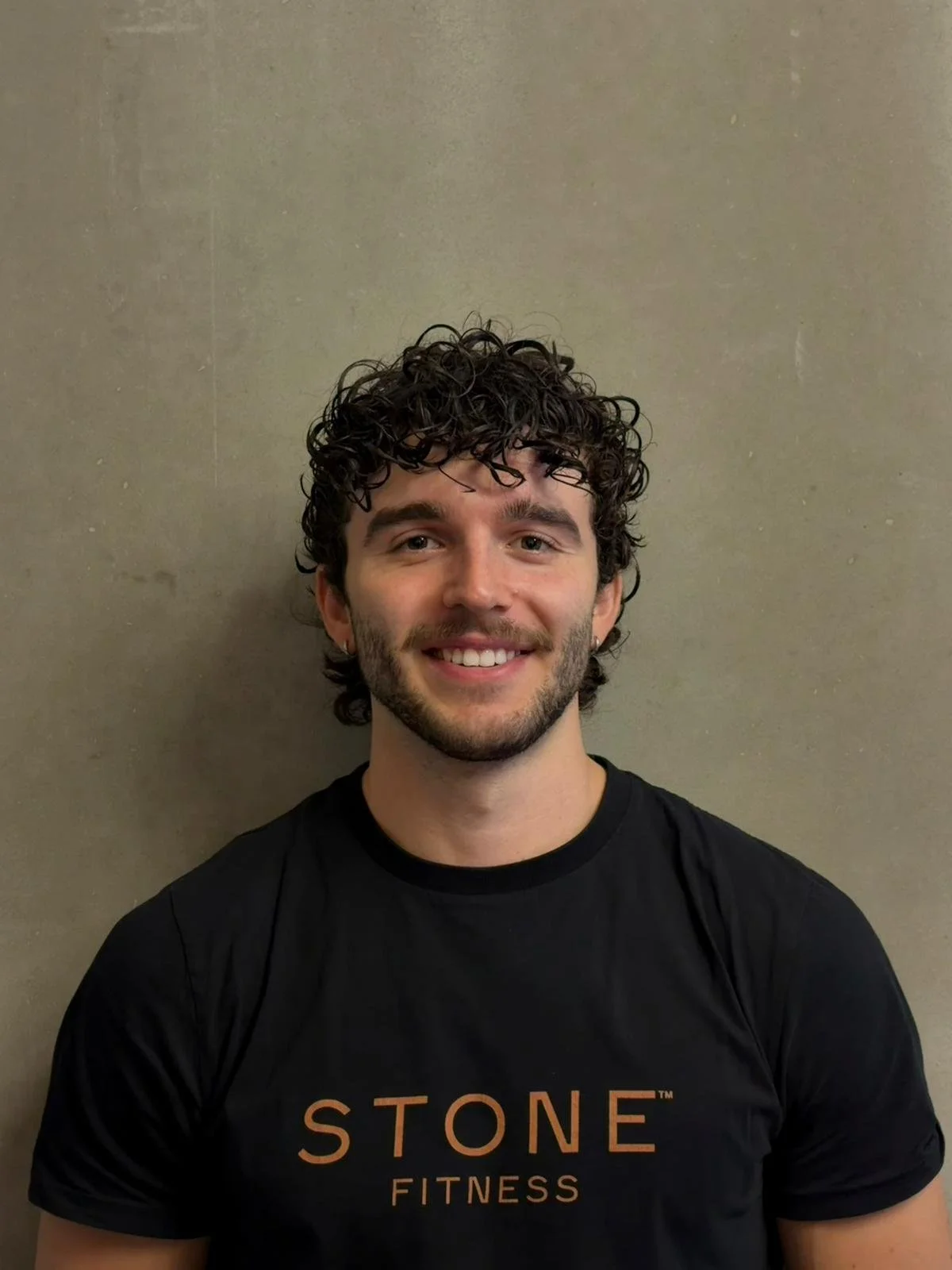 Portrait of a young man with curly hair and a beard, smiling, wearing a black shirt.
