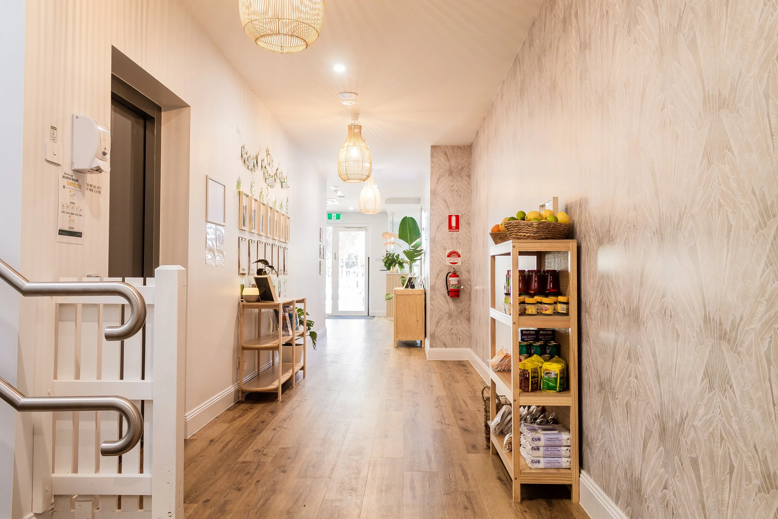 Bright indoor corridor with wooden flooring, light-colored walls, decorative wall art, and hanging pendant lights. Features a bookshelf with food items, potted plants, and a fire extinguisher near the entrance door at the end of the hall.