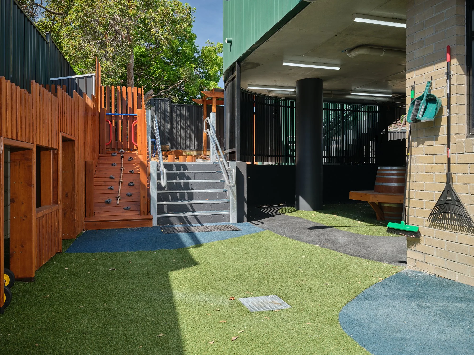 Child's outdoor play area with artificial grass, small climbing wall, stairs, and shed. Area includes a bench, cleaning tools hanging on the wall, and a small fenced space with trees overhead.