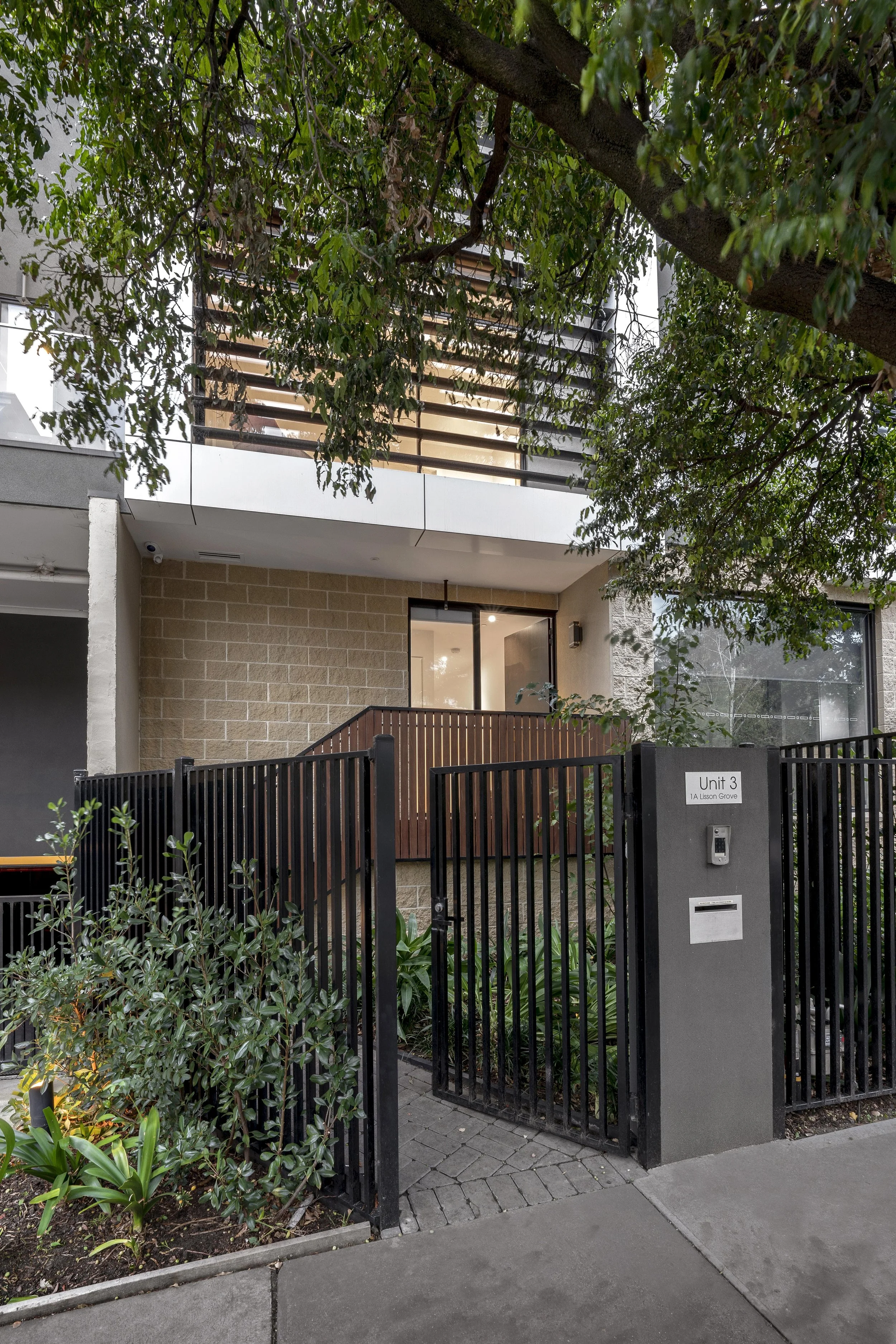 Modern residential apartment building with a gated entrance, greenery, and a large tree.