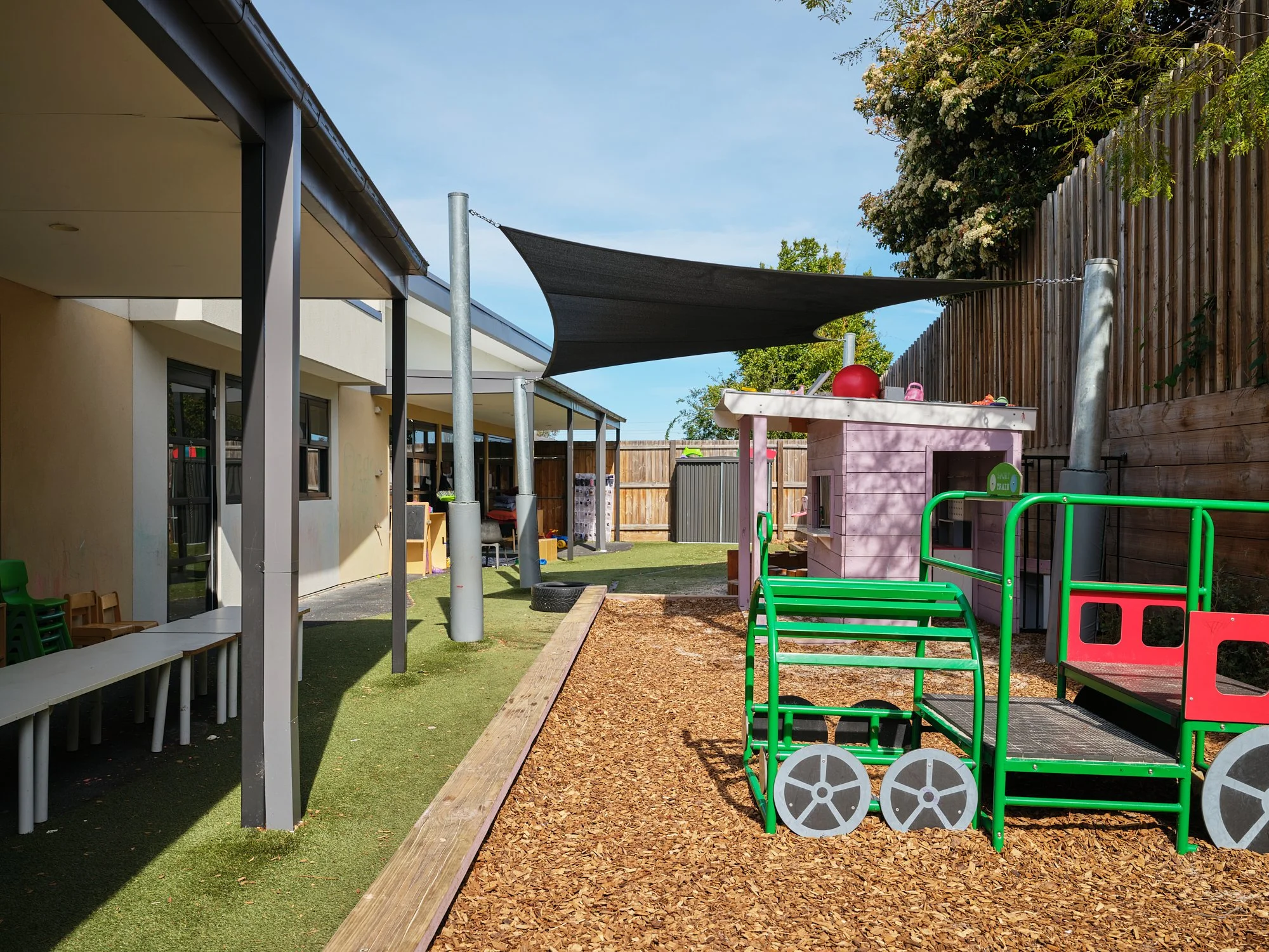 An outdoor playground area with a green metal play structure, a small pink playhouse, and a shaded area with a black canopy. There are wooden and metal fences, chairs, and a building with windows in the background.