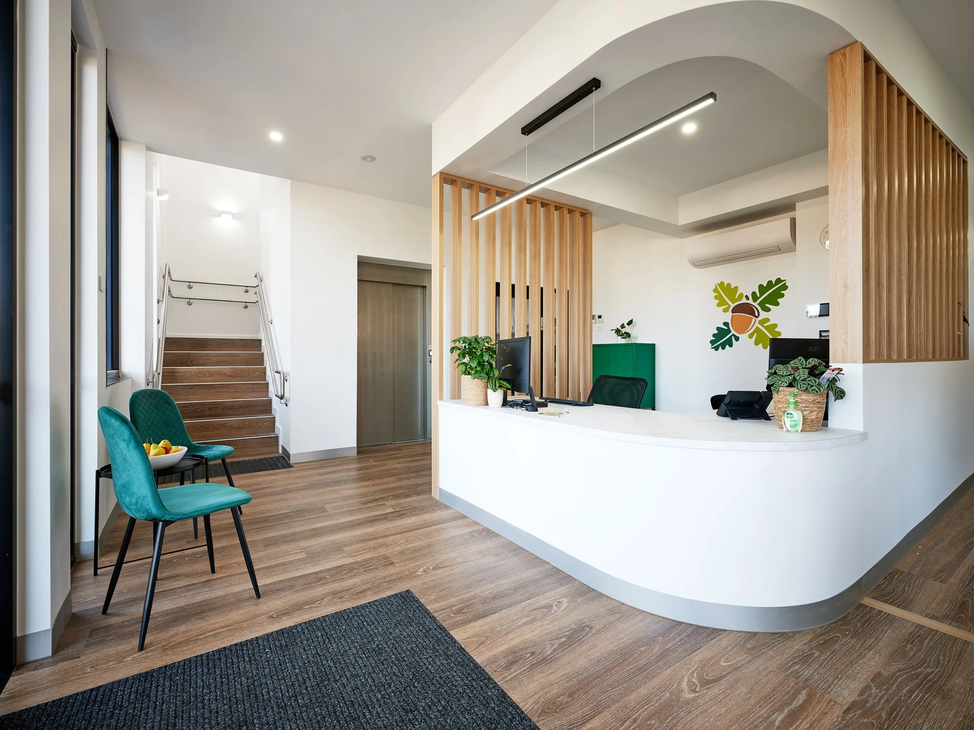 Modern reception area with white curved desk, wooden accents, green chairs, potted plants, and a wall decoration of an acorn with green leaves.