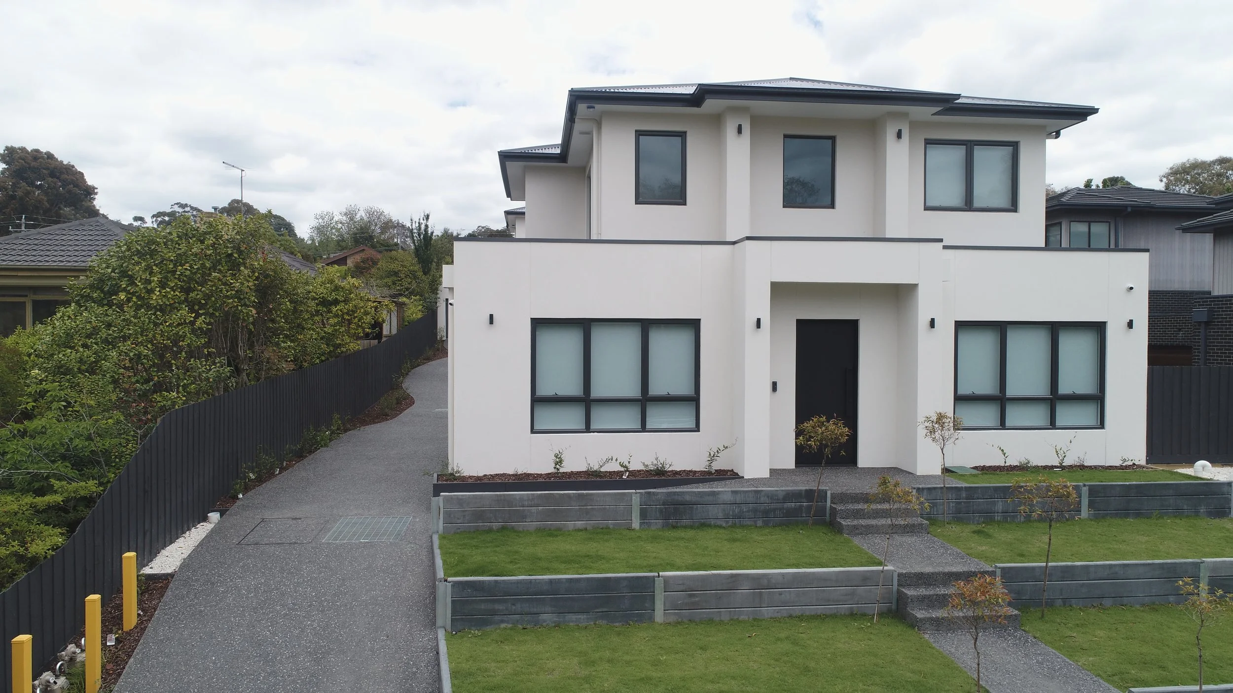 Modern two-story white house with large windows and a black front door, surrounded by a manicured lawn and small trees, with a winding driveway on the left.