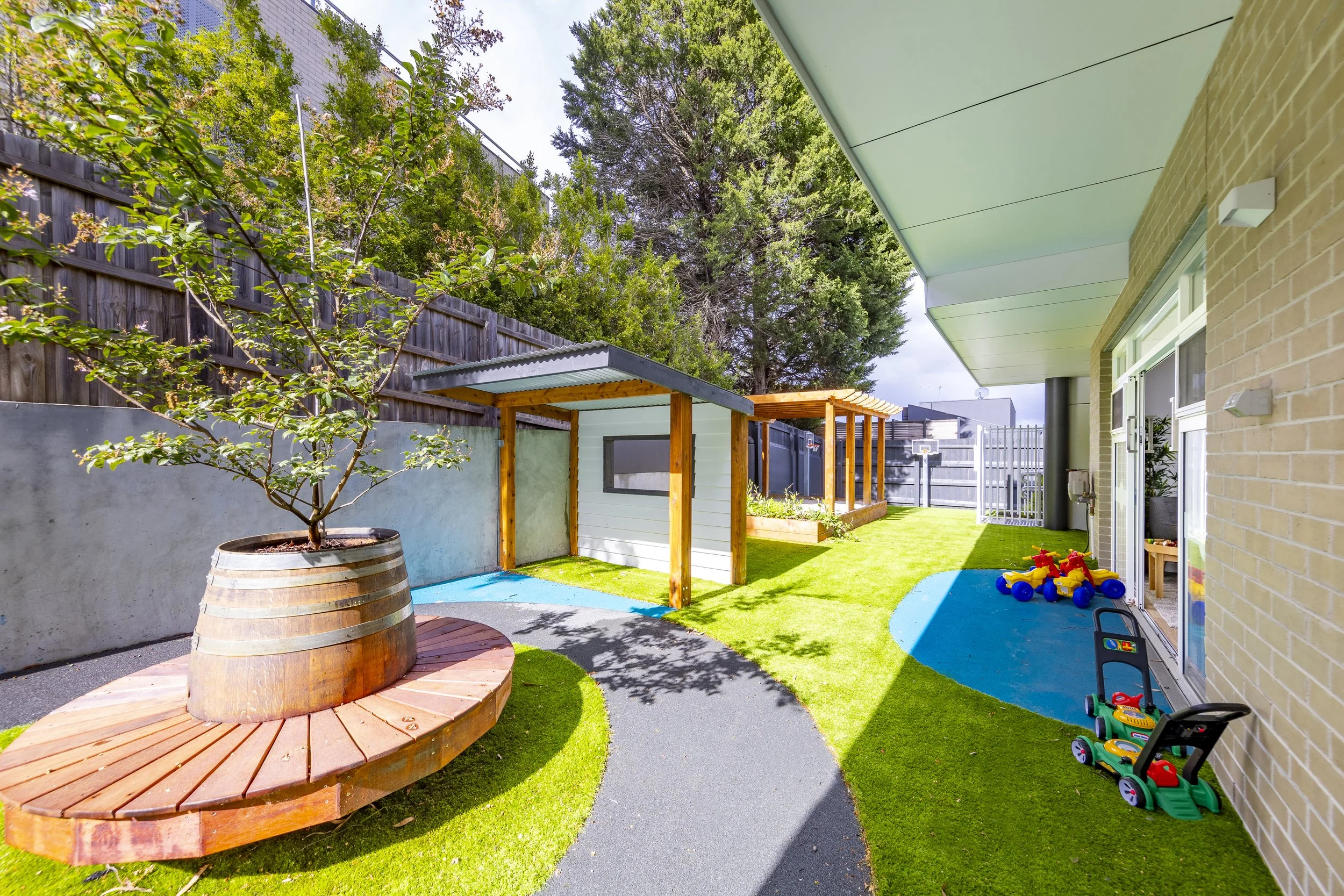 Backyard with artificial turf, a stone pathway, a large potted tree, a small playhouse, and children's toys near the house wall.