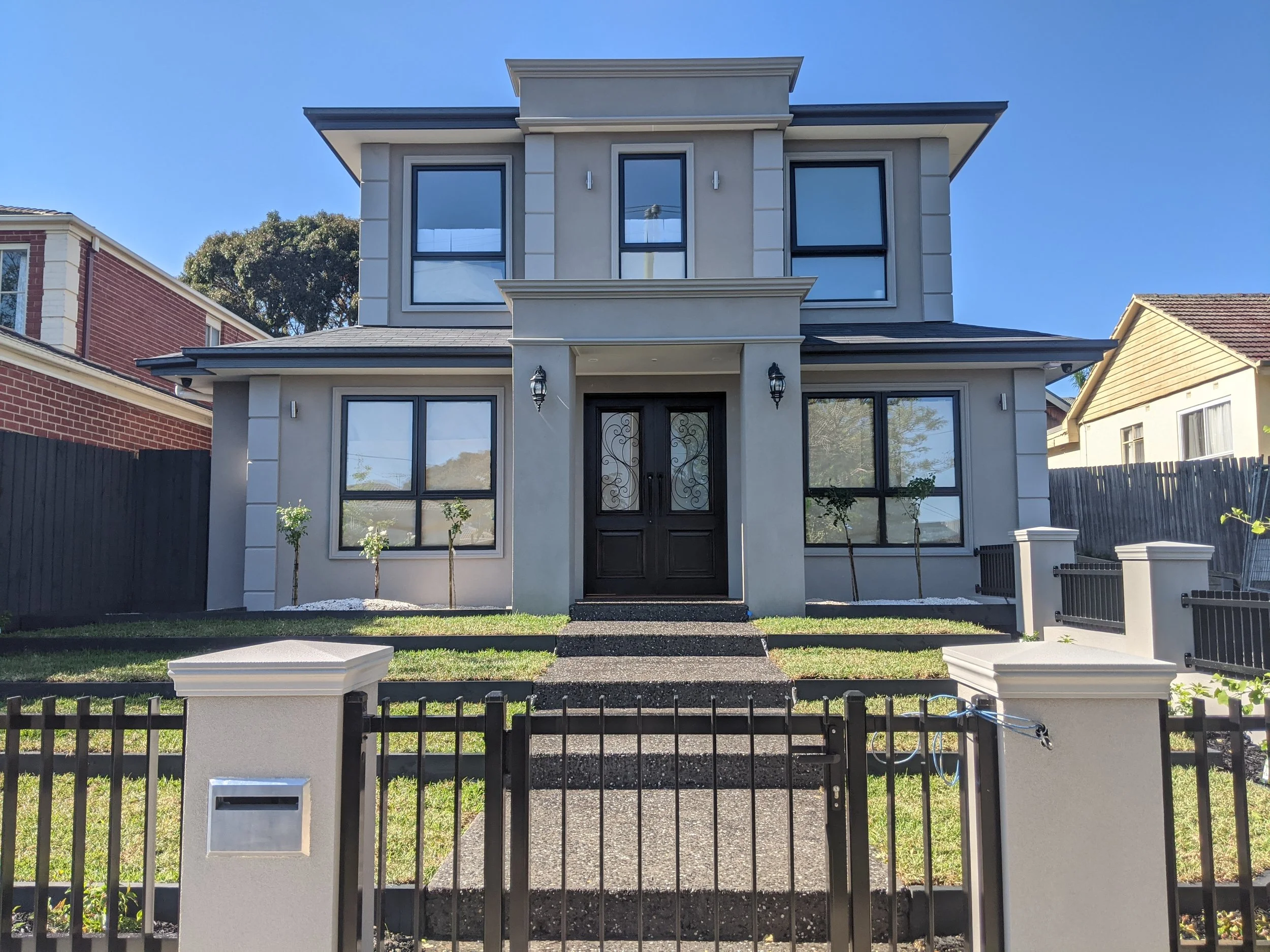 A modern two-story house with a front yard, black front door with decorative glass, black gate, and steps leading to the entrance. The house has large windows, some with curtains, and is flanked by smaller trees planted in the yard.