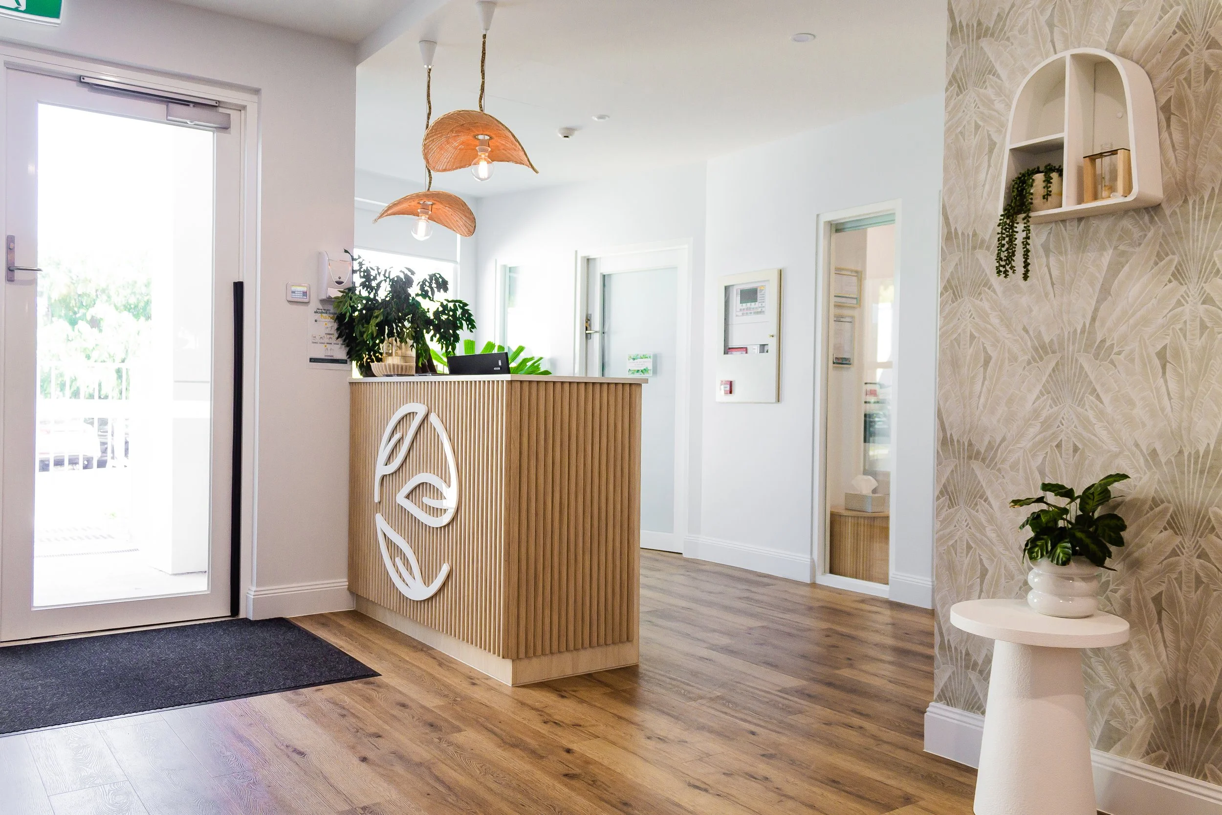 Modern front desk area with wooden counter, indoor plants, two hanging pendant lights, white walls, and decorative wallpaper with leaves pattern.