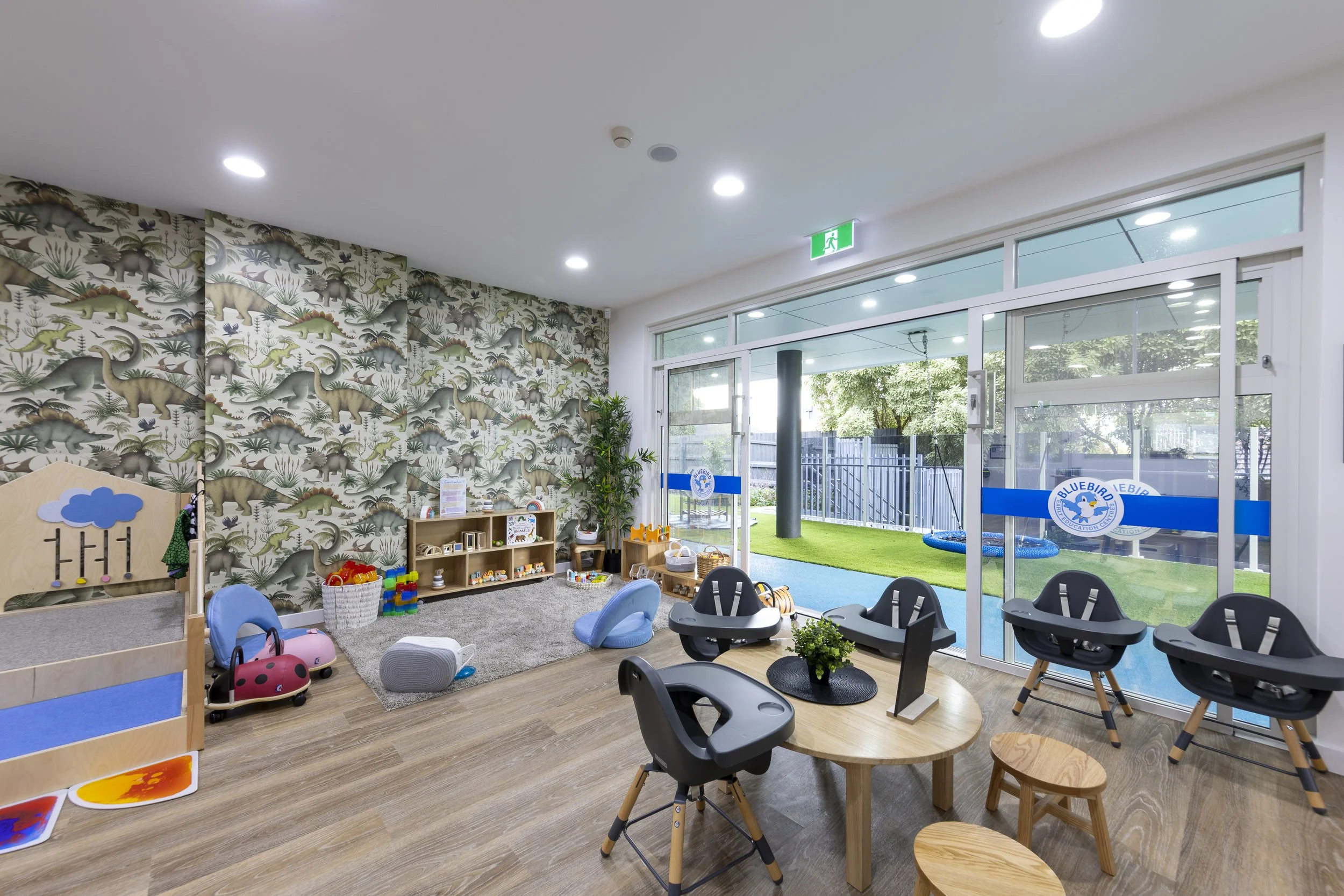 Indoor children's playroom with toys, small chairs, and a window showing outdoor play area with grass and play equipment.