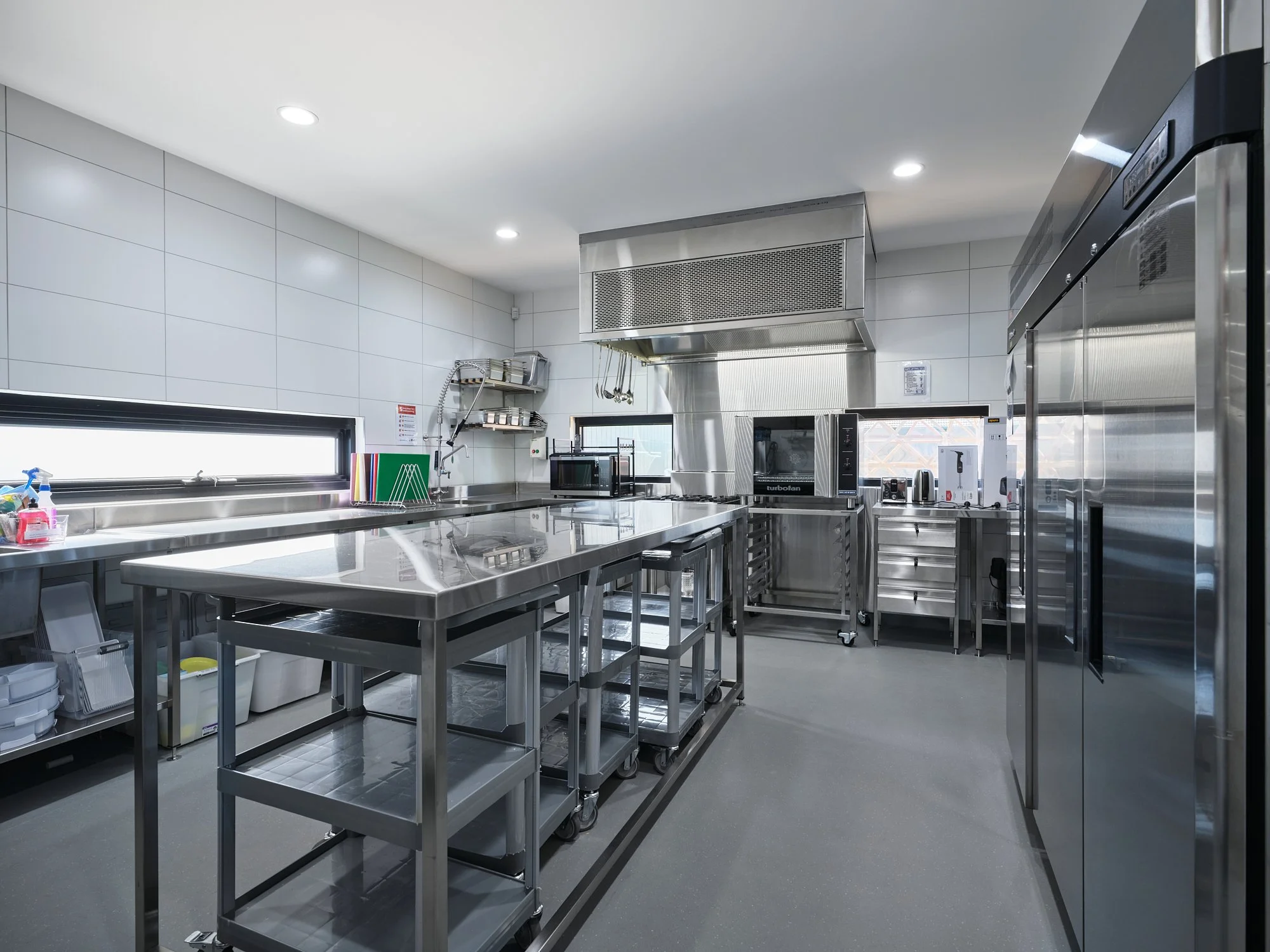 Sterile commercial kitchen with stainless steel counters, shelving, and appliances, illuminated by overhead lighting and a horizontal window.