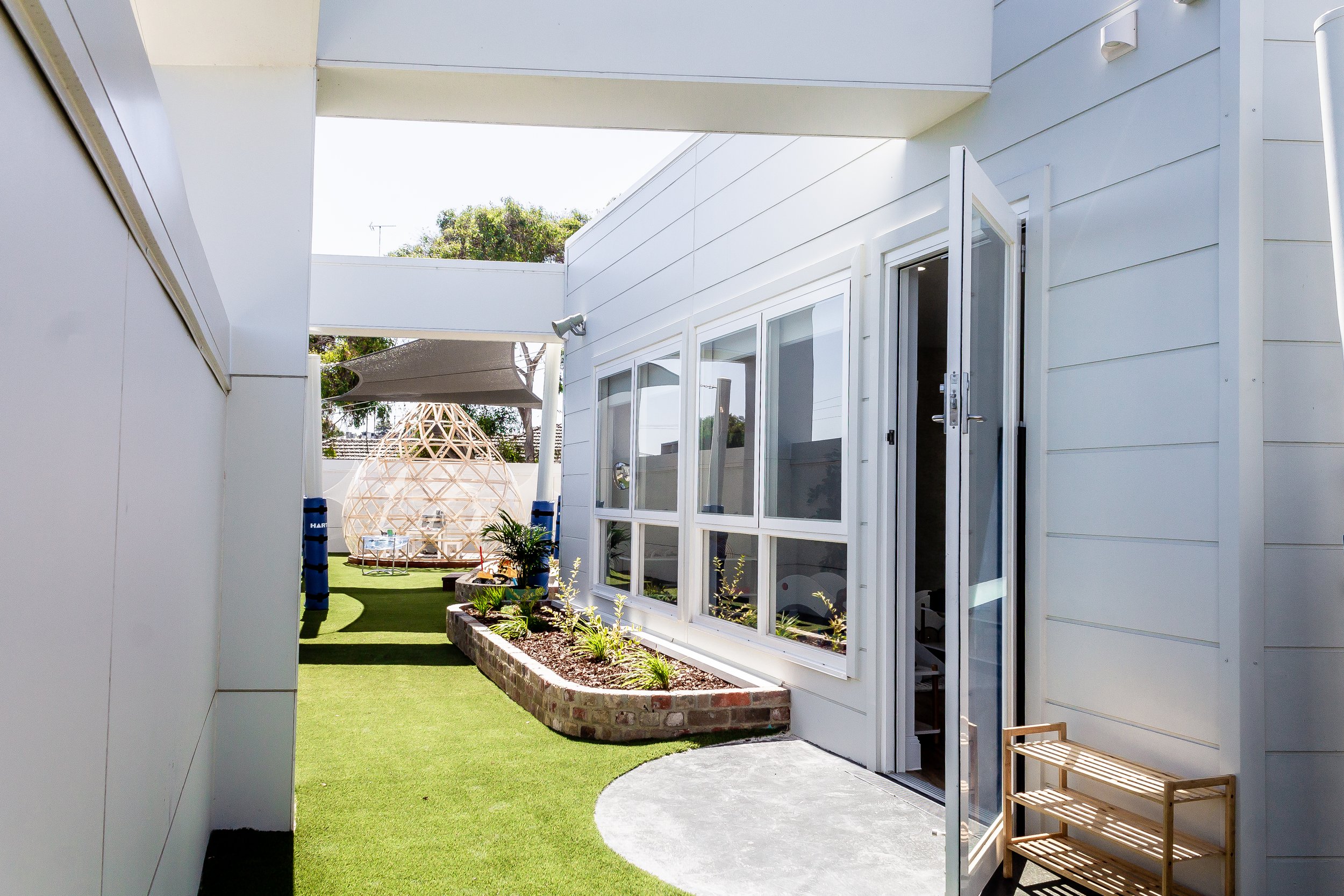View of a modern white house's backyard with a sliding glass door, exposed brick planter, green artificial grass, and a large wooden geodesic dome structure in the background.
