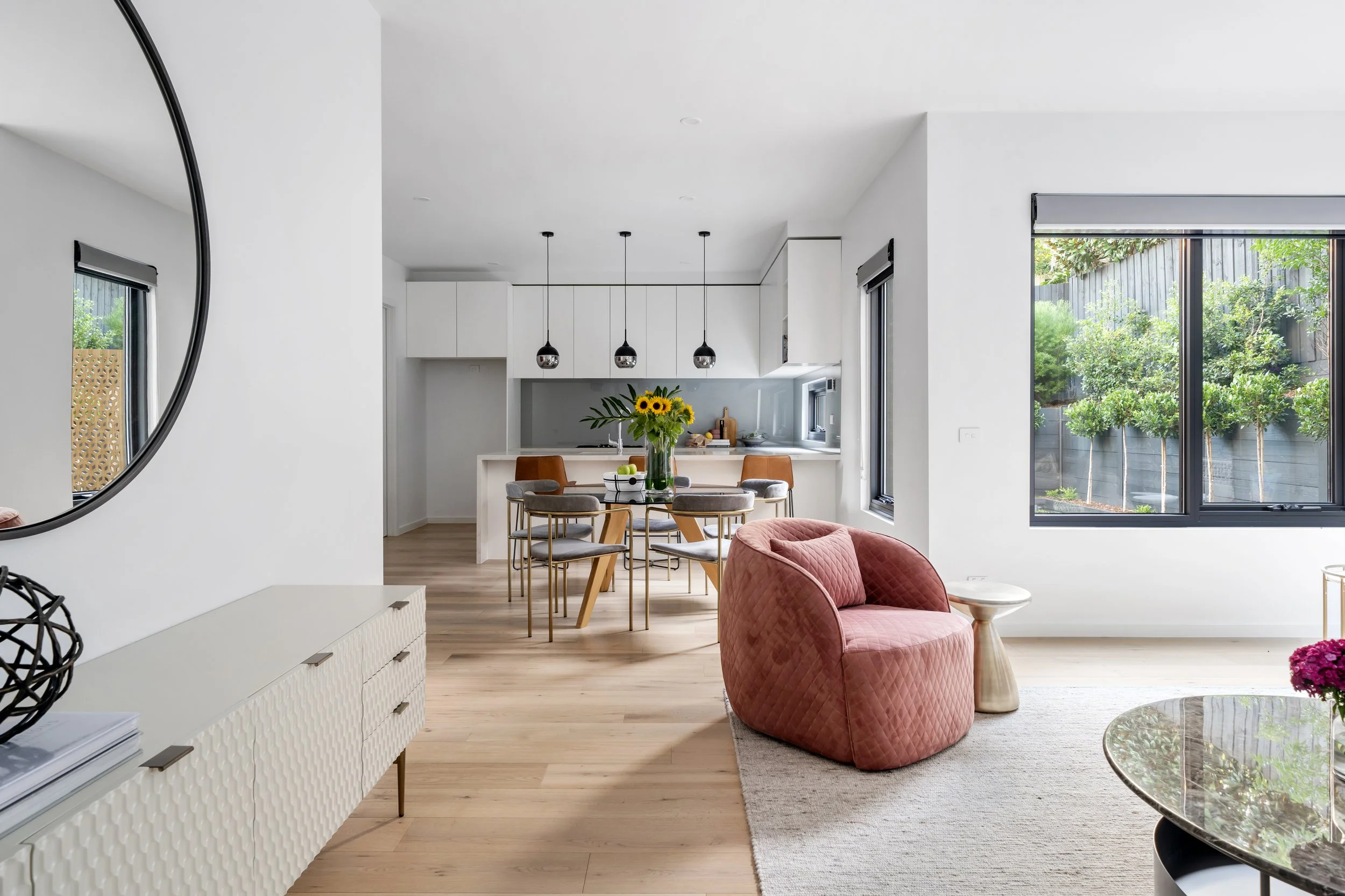Modern living room with pink quilted armchair, glass coffee table, light wood flooring, white wall, and large window showing greenery outside, adjacent to an open kitchen and dining area with sunflowers on the table.