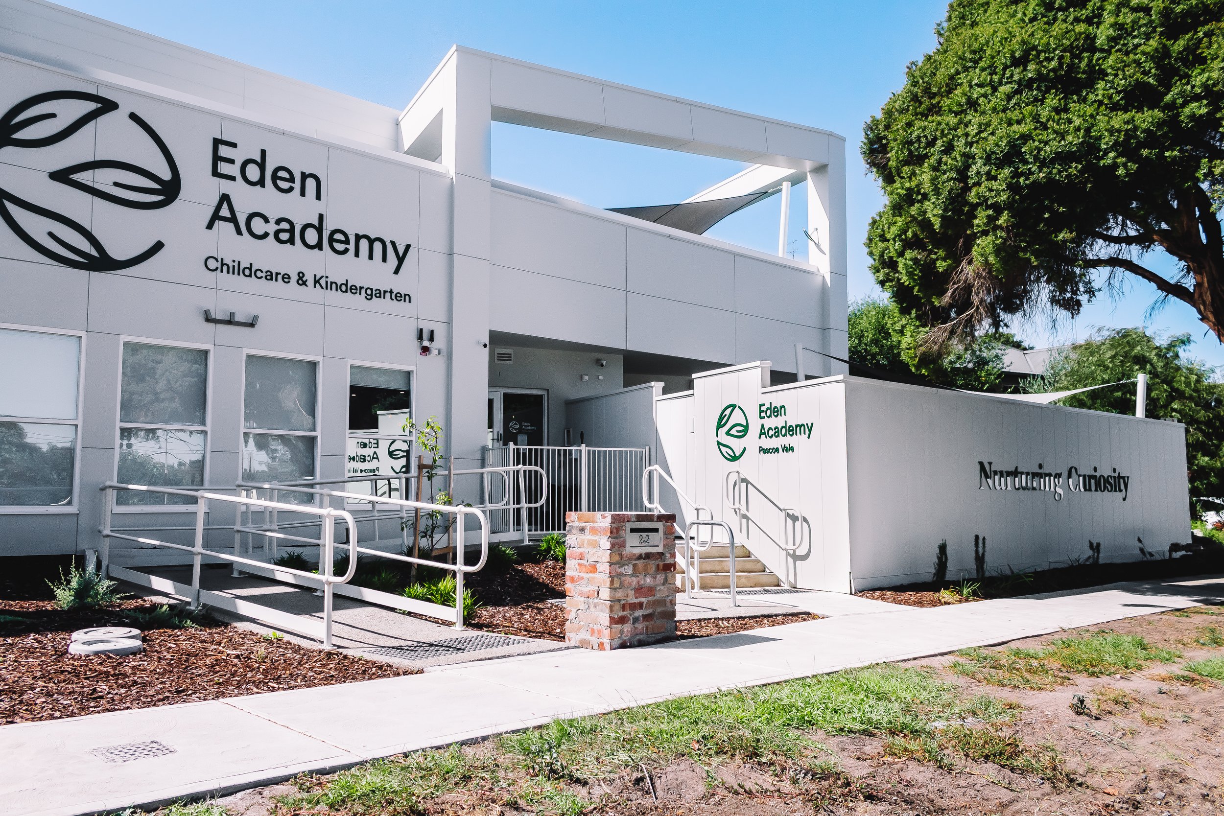 Exterior of Eden Academy childcare and kindergarten center with white modern building, logo with leaves, and slogan 'Nurturing Curiosity,' surrounded by trees and sidewalk in a sunny day.