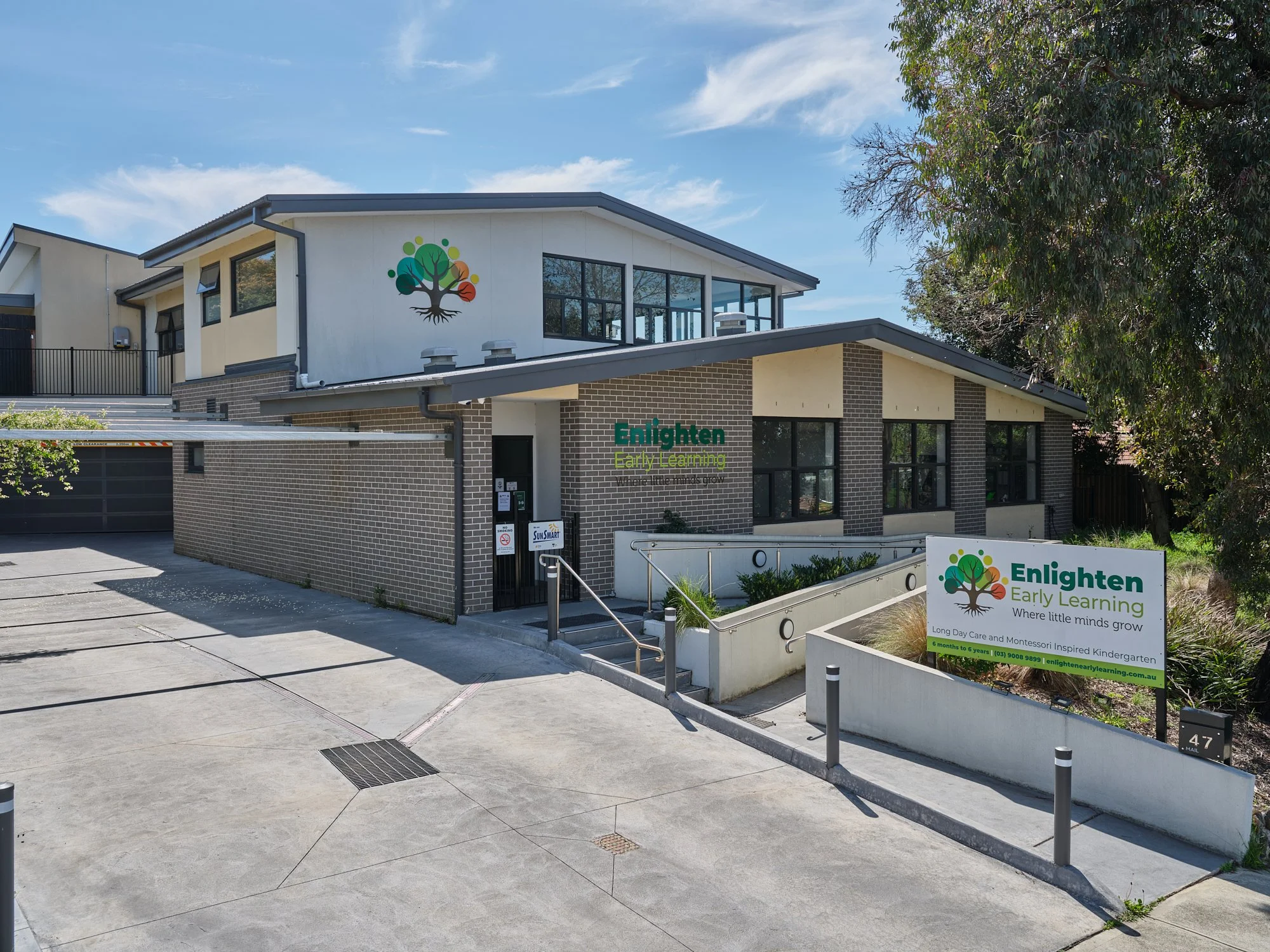 Exterior of Enlighten Early Learning center with the sign and a building featuring large windows, brick and white walls, a ramp for accessibility, and a colorful tree logo on the upper wall.