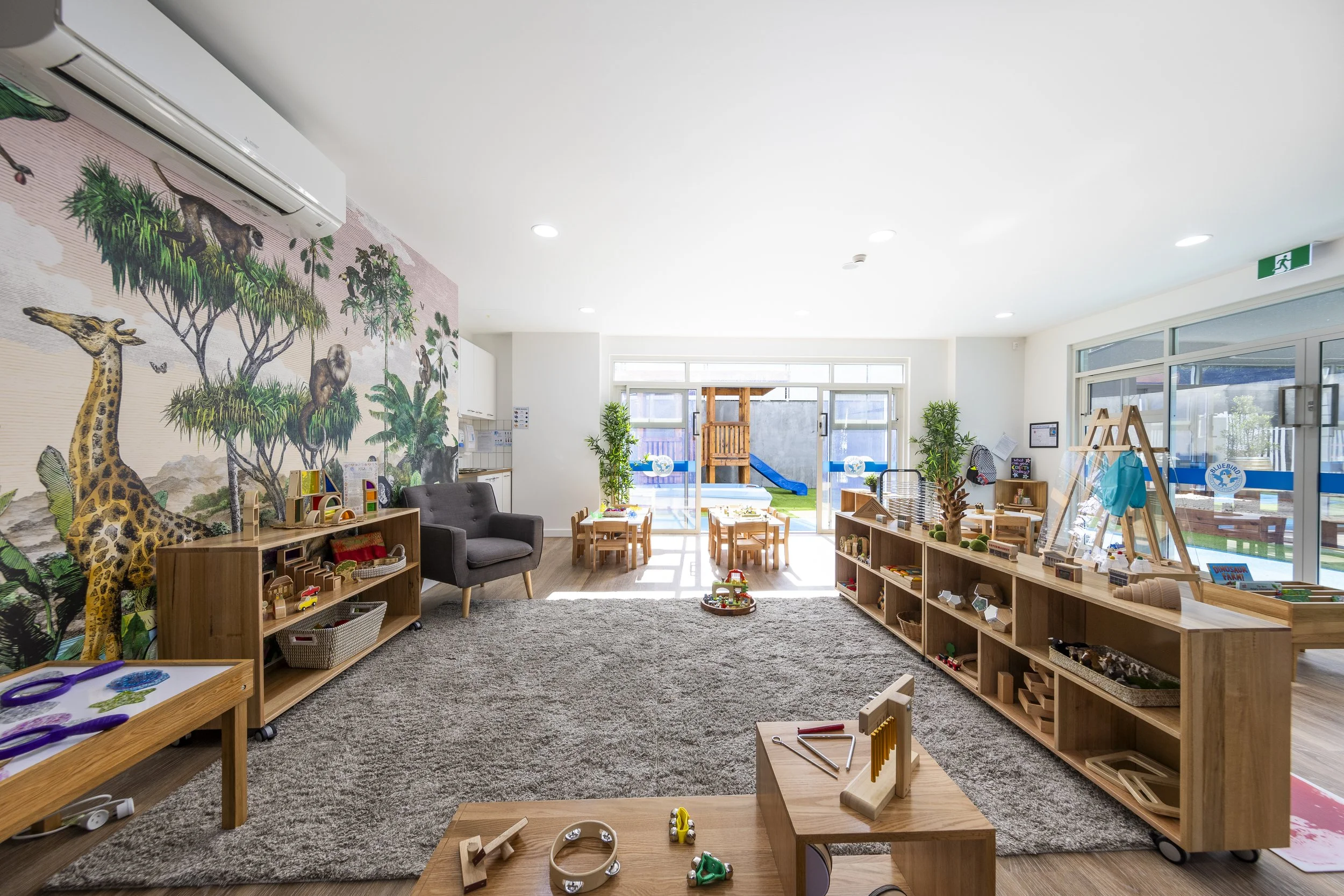 Bright, colorful children's playroom with toys, wooden furniture, a jungle-themed mural, and a sliding glass door leading to an outdoor play area with a slide.