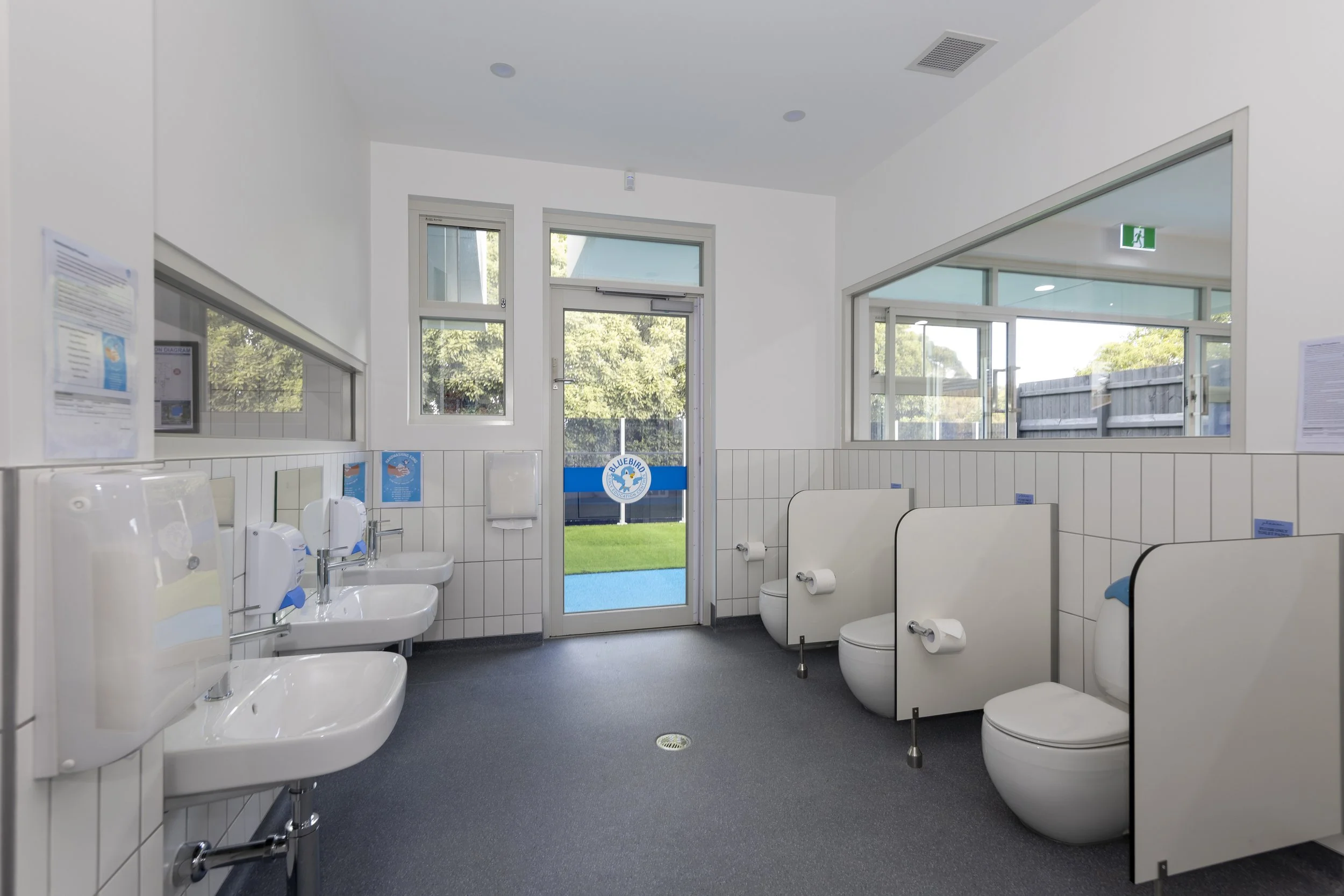 Public restroom with three sinks on the left, three toilets with partition privacy panels on the right, large windows allowing natural light, and a door with a blue and white logo that says 'Bluebird' at the back. The walls are white with tiled lower