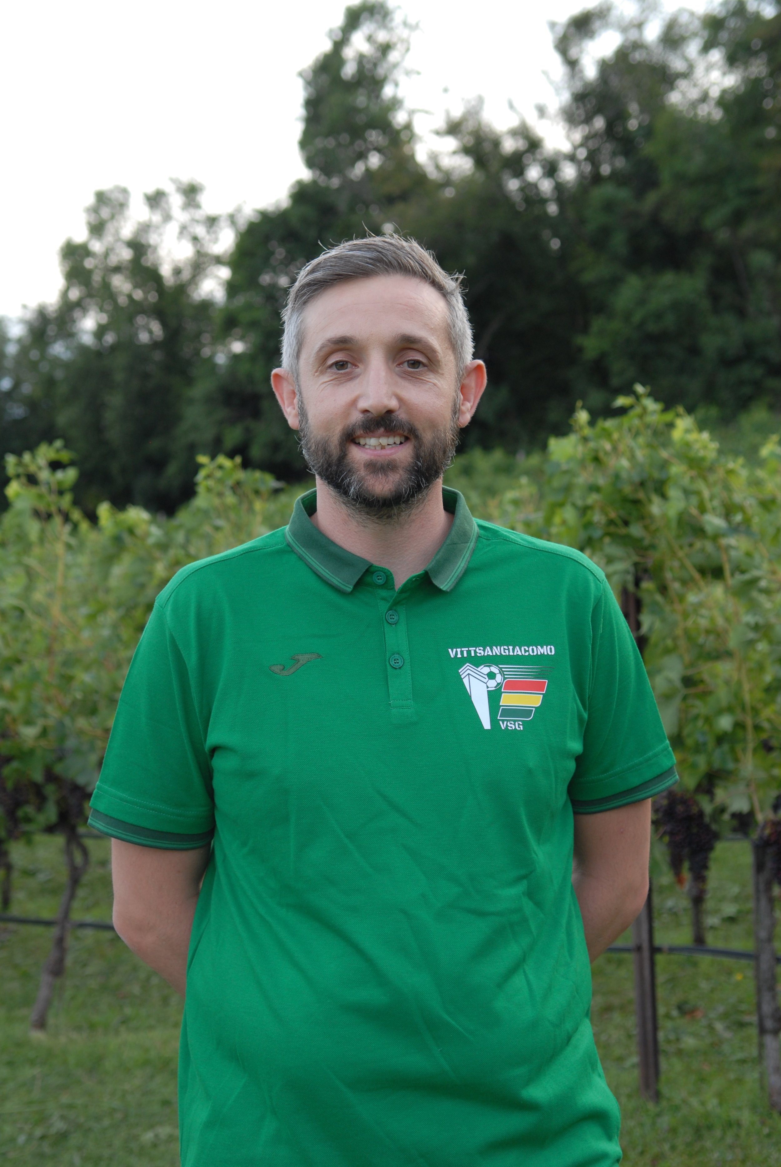 Uomo con capelli e barba, indossa una maglietta verde con logo e scritte, si trova in un campo con viti e alberi sullo sfondo.