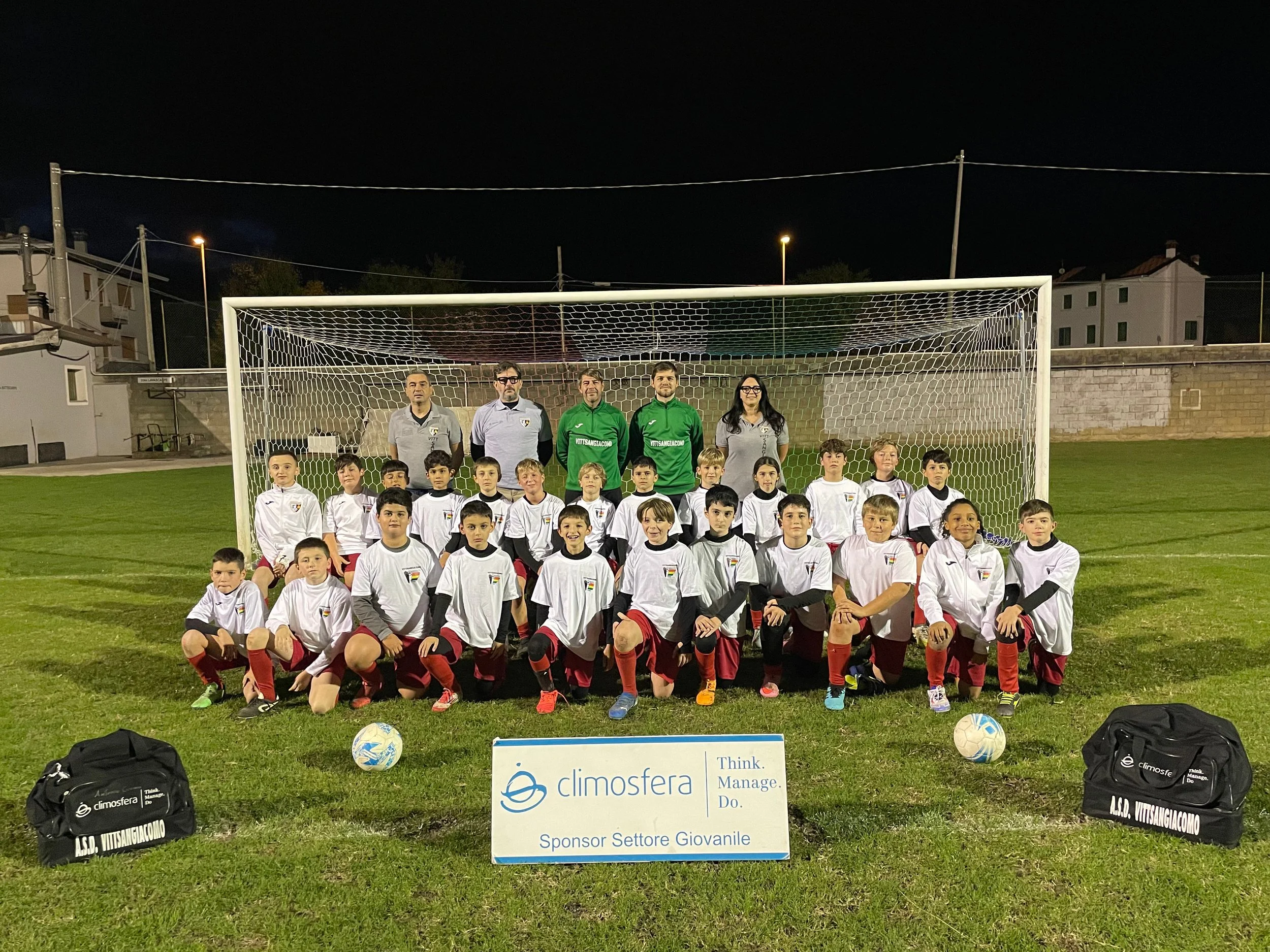 Gruppo di bambini calciatori con allenatori in divise sportive davanti a un goal durante una partita di notte. In primo piano ci sono due borsoni e un cartello con il logo