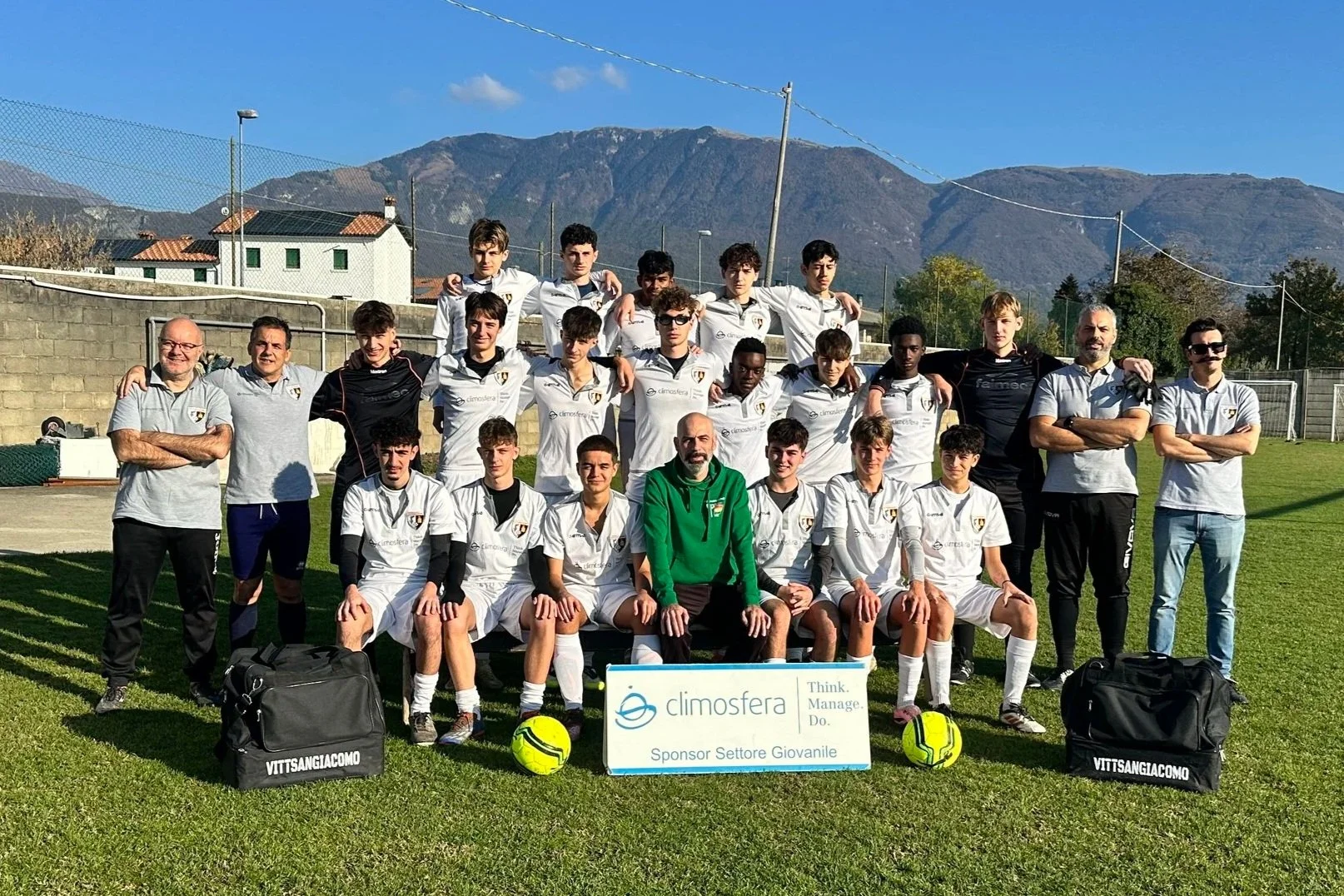Gruppo di giovani calciatori e allenatori sulla linea di centrocampo di un campo da calcio, con montagne sullo sfondo, in un giorno soleggiato.