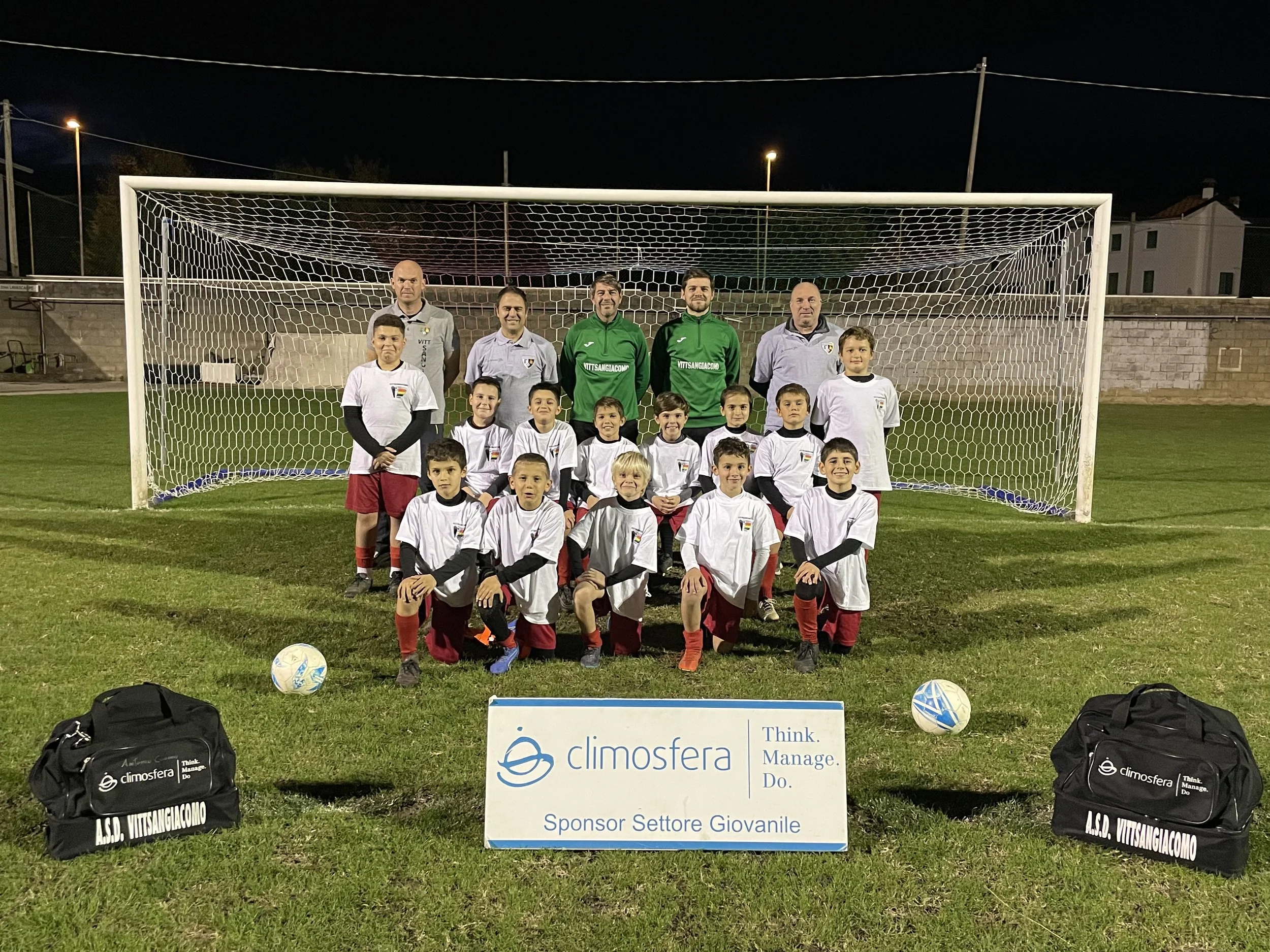 Gruppo di bambini e adulti di calcio posizionati davanti a una porta da calcio, con palloni e borse sportive sul prato, in una serata diurne o notturna.