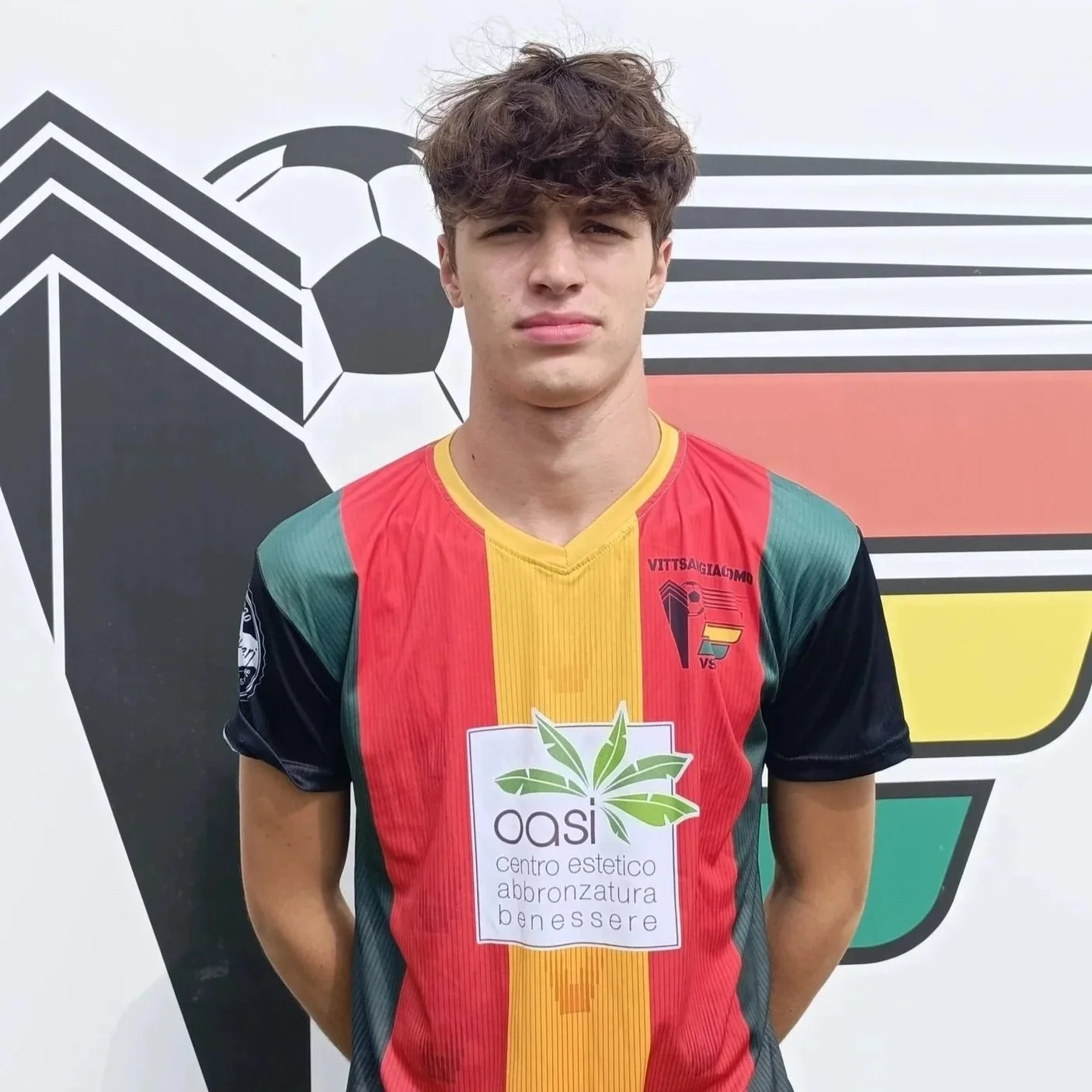 Ragazzo con maglietta di calcio colorata in rosso, giallo e verde, con logo di sponsorizzazione e sfondo grafico con figure geometriche e una palla da calcio.
