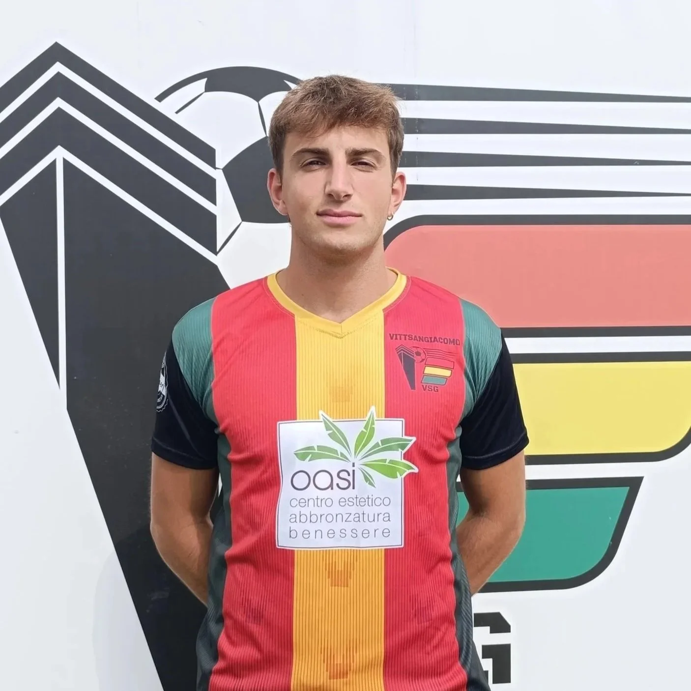 Giovane uomo che indossa una maglia da calcio rossa, gialla e verde, davanti a uno sfondo con un logo astratto e un disegno di calcio
