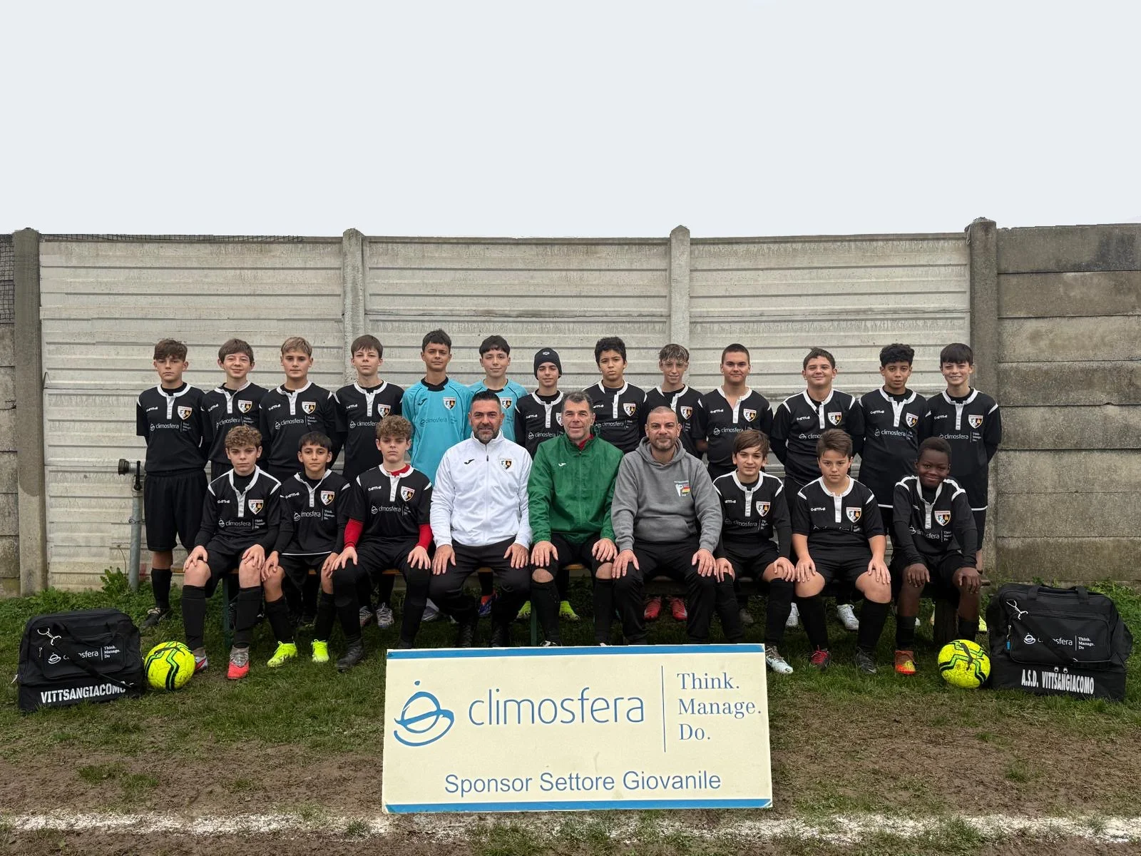 Gruppo di giovani calciatori con tre allenatori in piedi e seduti davanti a una recinzione di cemento, con palloni da calcio e borse sportive ai lati, in posa per una foto di squadra.