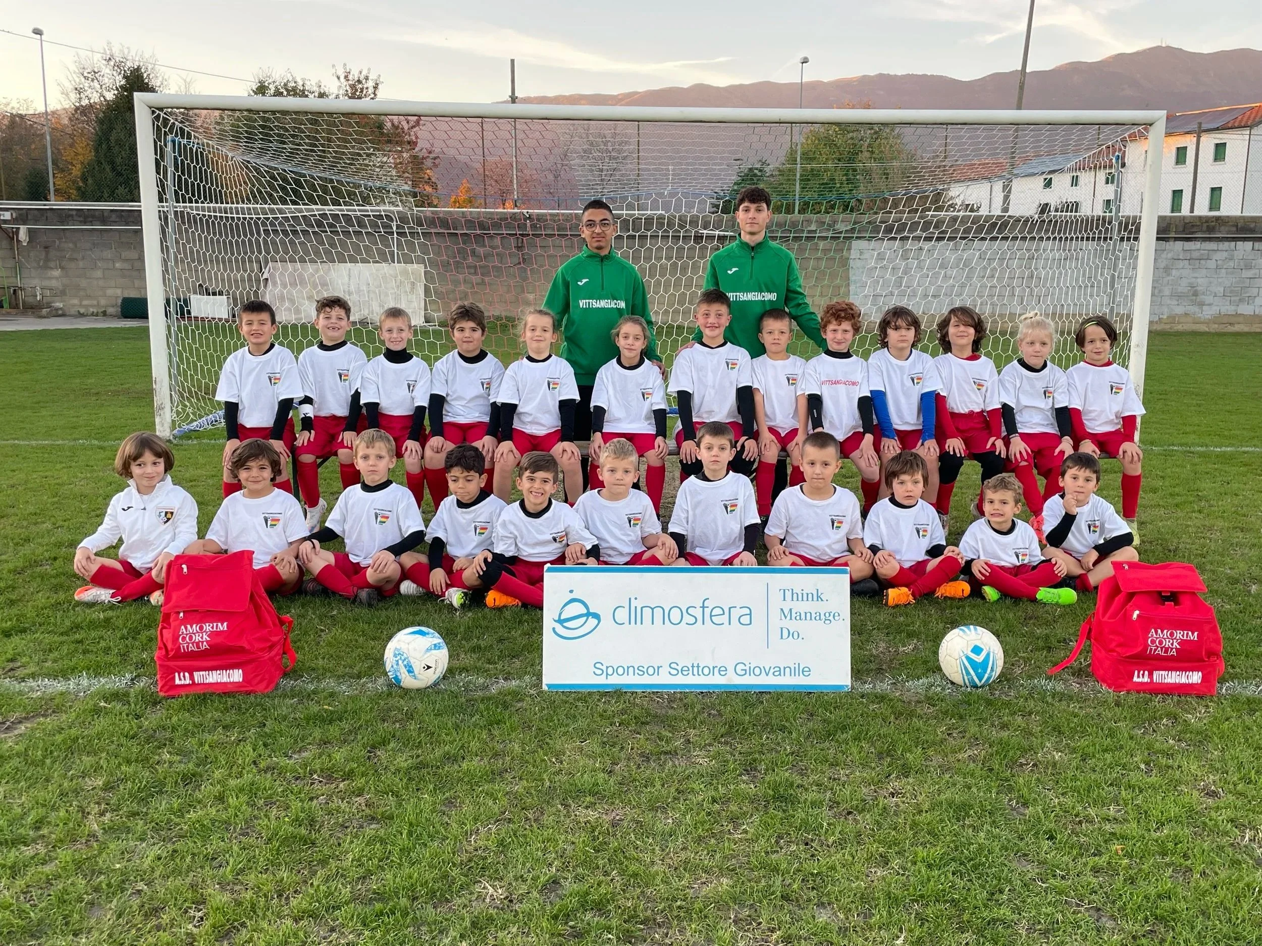 Gruppo di bambini calciatori in divise bianche e rosse, accompagnati da due adulti in maglietta verde, posano davanti a un campo di calcio con una rete. Ci sono due palloni e uno striscione con il logo di Climosfera e testi in italiano.