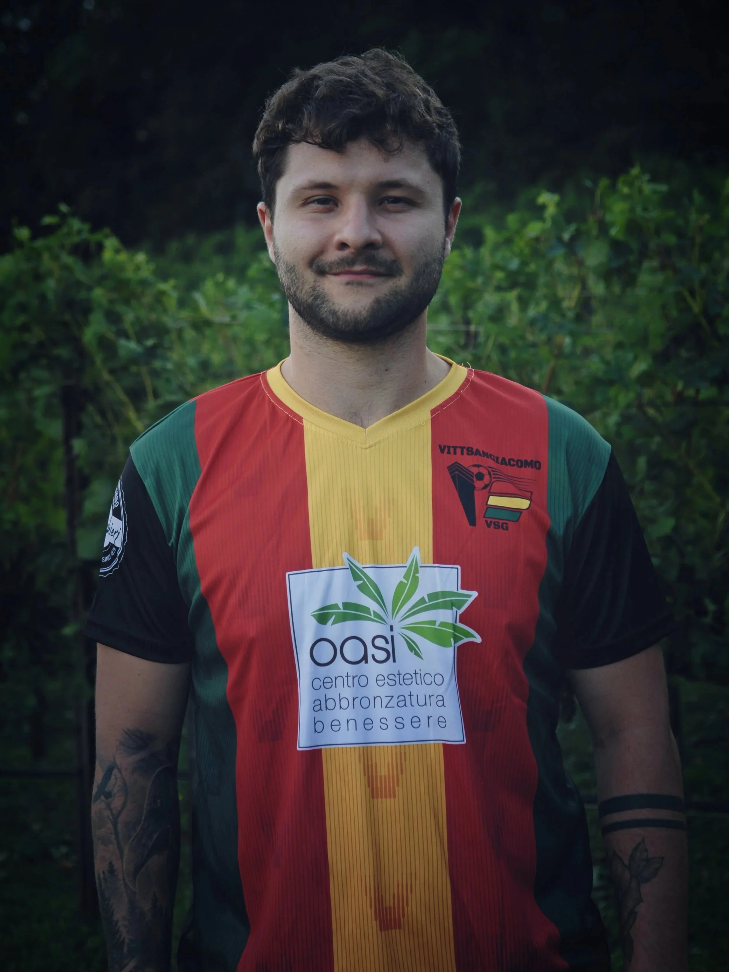 Giovane uomo con barba e tatuaggi, indossa una maglietta da calcio con strisce verdi, rosse e gialle, sullo sfondo un'area verde