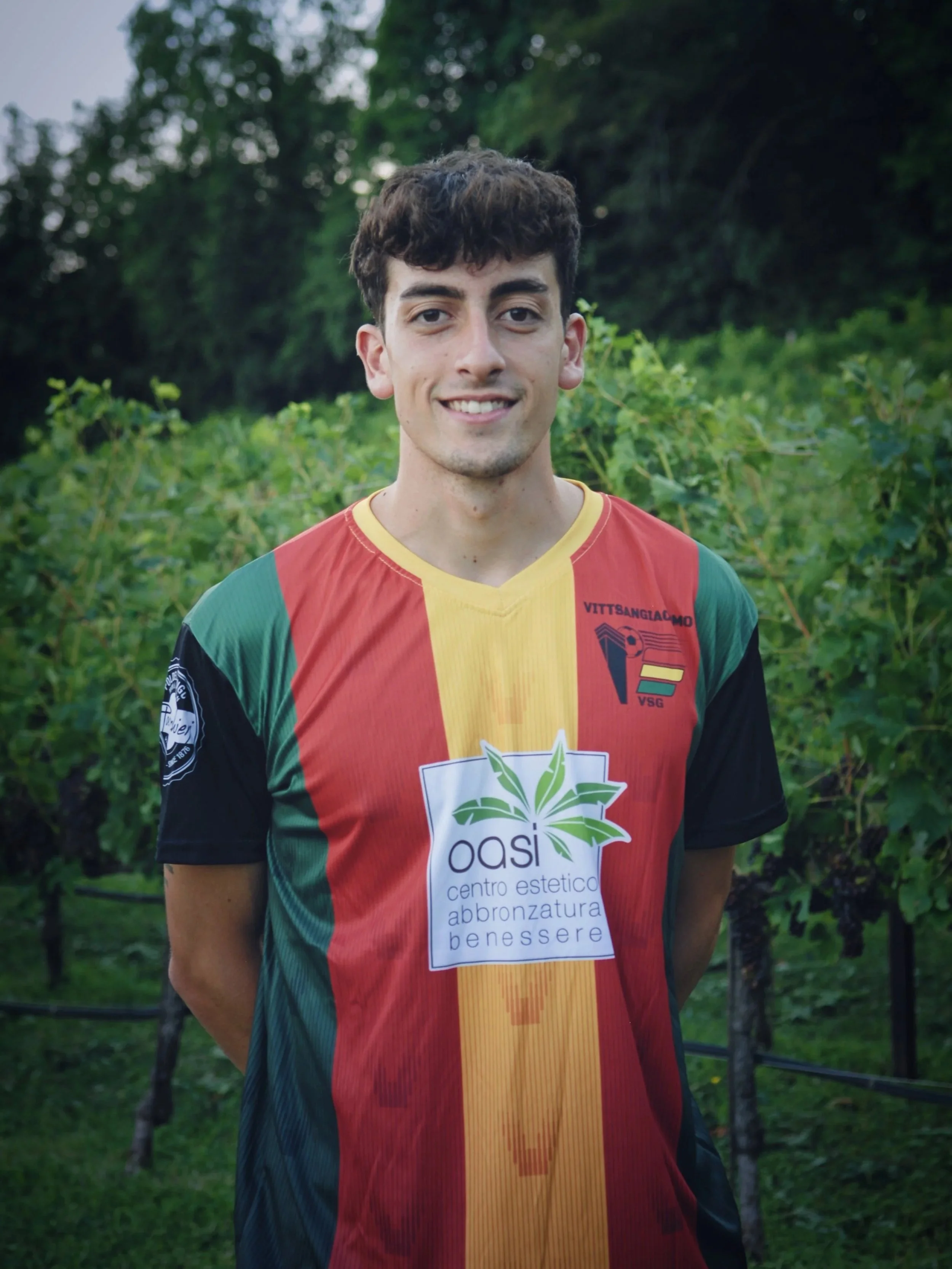 Giovane calciatore sorridente con maglietta a righe di colori rosso, giallo, verde e nero, in piedi all'aperto tra piante e alberi.