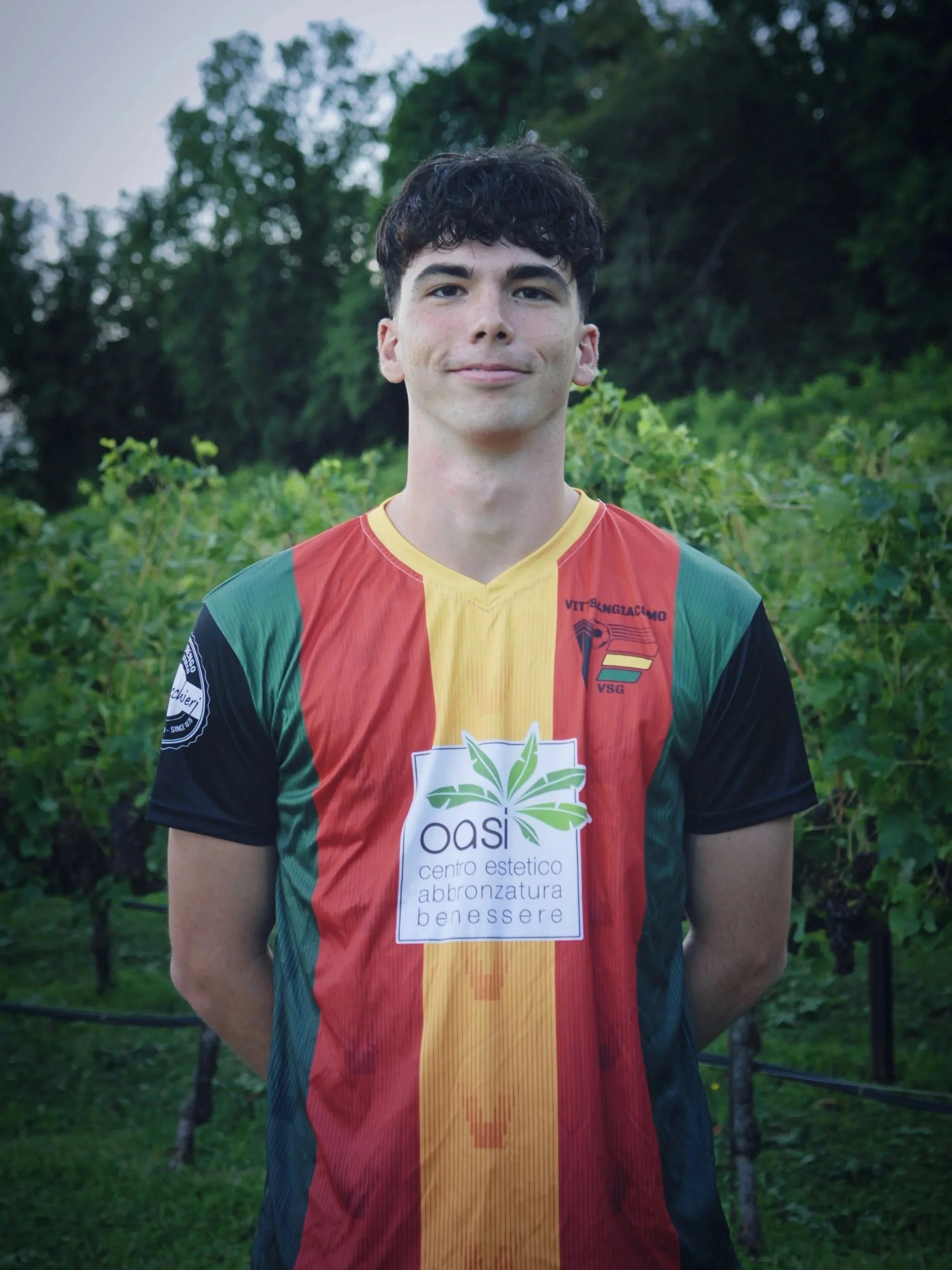 Giovane uomo con capelli ricci scuri indossa una maglietta da calcio a strisce colorate rosso, giallo, verde e nero. È in piedi in un ambiente naturale con vegetazione verde alle spalle.