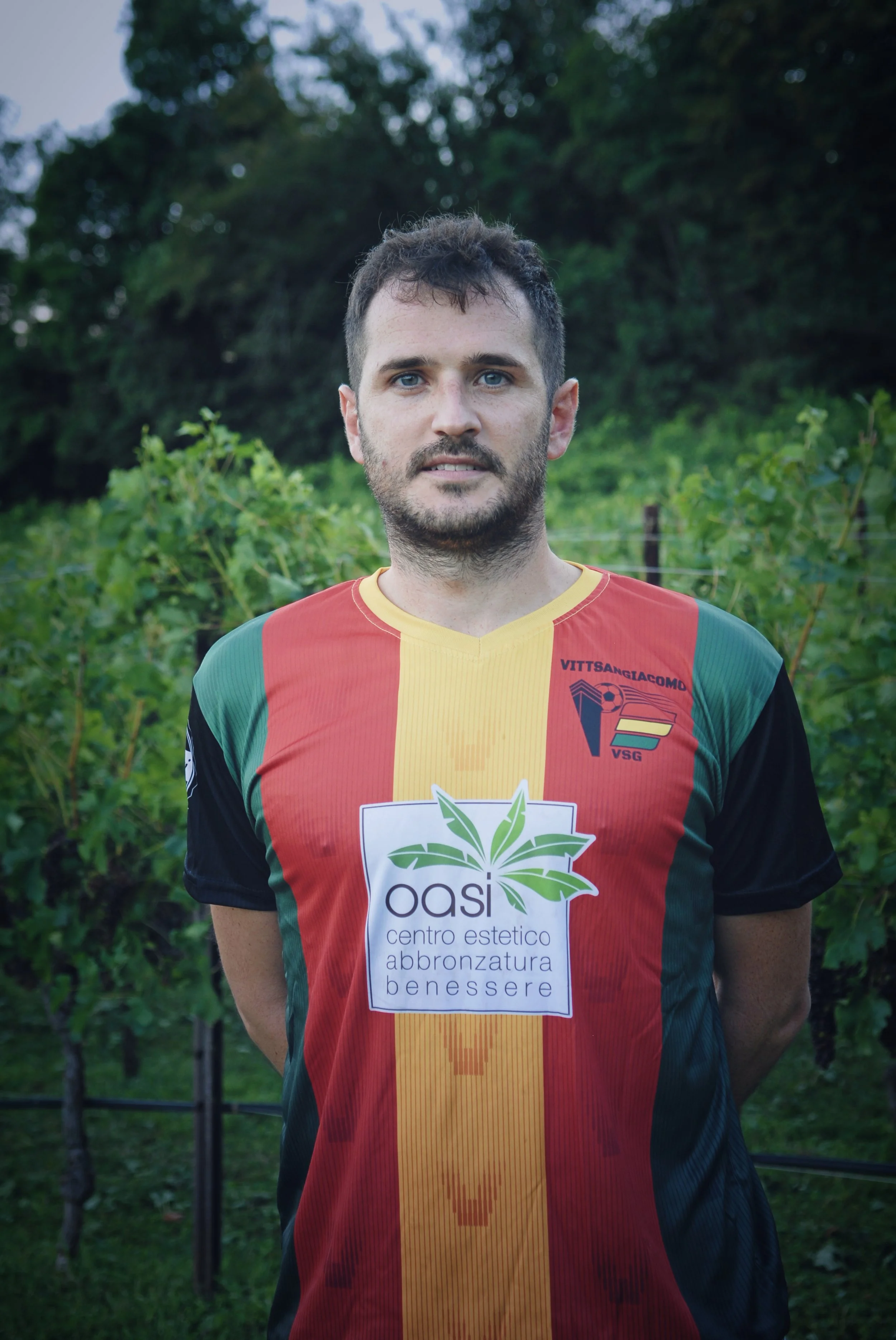 Uomo con capelli castani, barba e occhi chiari indossa una maglietta da calcio con i colori rosso, giallo e verde, in piedi in un'area verde con arbusti e alberi sullo sfondo.