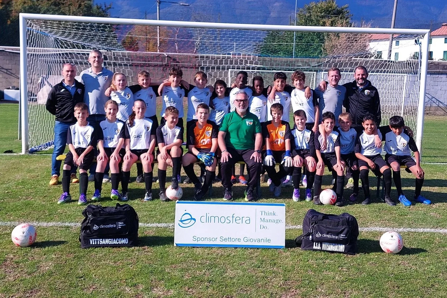 Gruppo di bambini di calcio con i loro allenatori in campo, posizionati davanti a una porta da calcio, con palloni e una cartellina con sponsorizzazioni, in una giornata di sole.