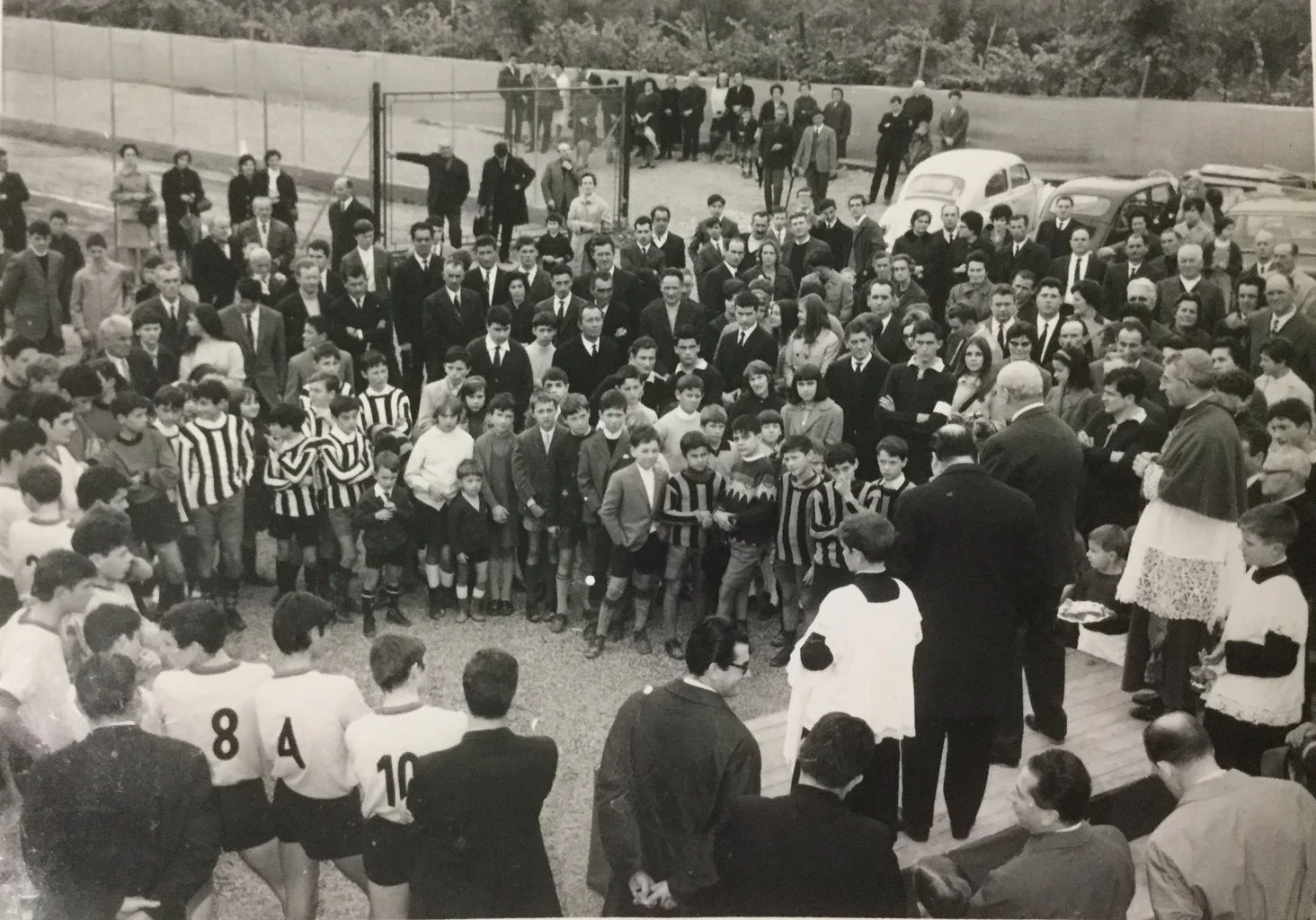 Un gruppo di persone, inclusi bambini e adulti, si trovano in cerchio su un campo, forse in occasione di un evento simbolico o commemorativo, con vari veicoli d'epoca e una recinzione sullo sfondo.