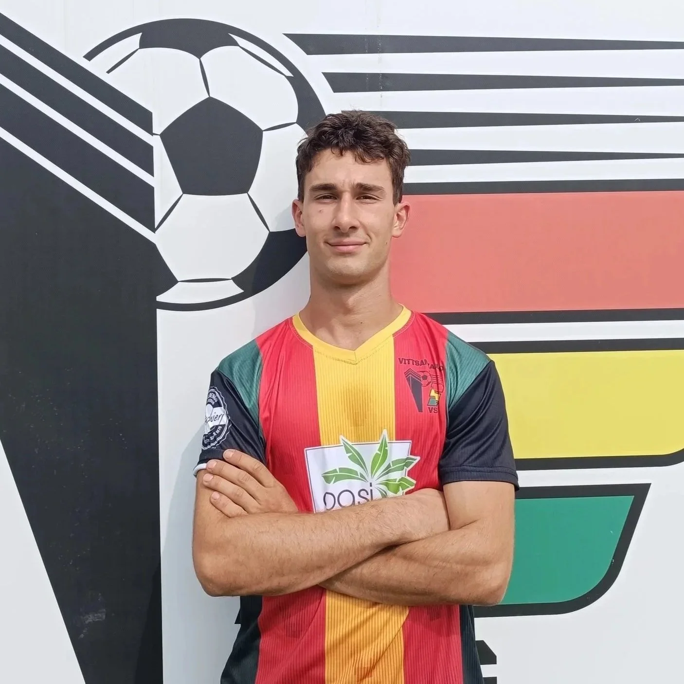 Giovane uomo in maglietta da calcio con colori giallo, rosso e verde, con braccia incrociate, posa davanti a un murale con un pallone da calcio e una bandiera.