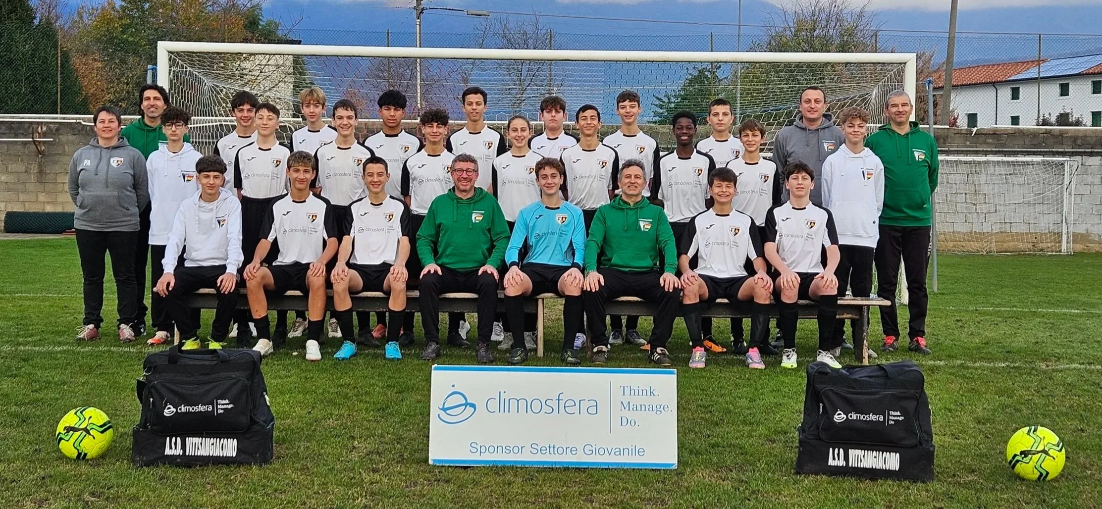 Gruppo di giovani calciatori e allenatori su un campo da calcio in posa per una foto di squadra, con due palloni da calcio gialli e due borse sportive in primo piano. Alla loro base un cartellone con logo e testo sponsorizzazione.
