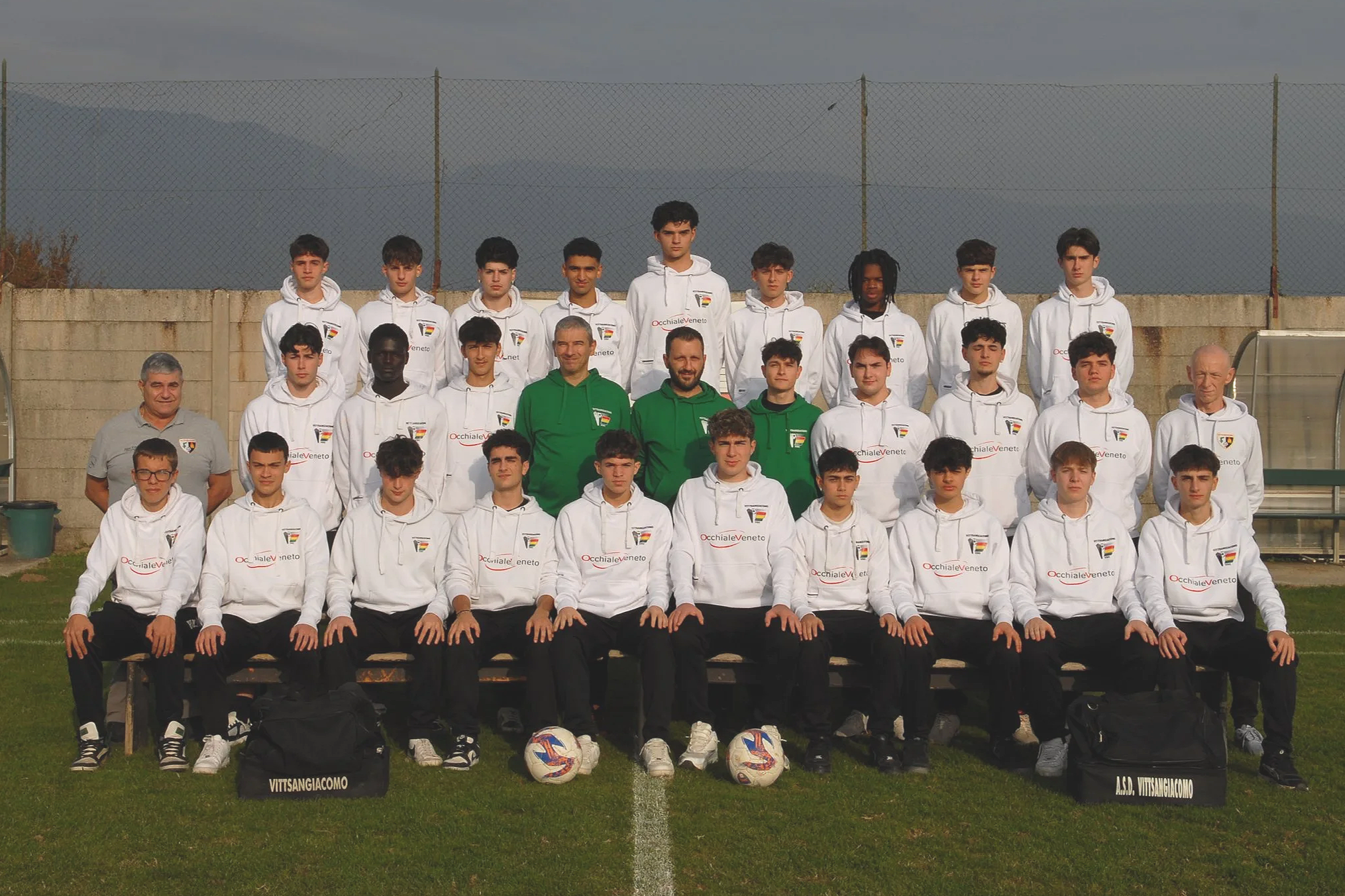 Gruppo di giovani calciatori e allenatori posizionati in campo, con alcuni in piedi sullo sfondo e altri seduti davanti, tutti vestiti con cappotti o felpe bianche con logo della squadra e sponsor, in uno stadio o campo sportivo, durante una sessione fotografica.