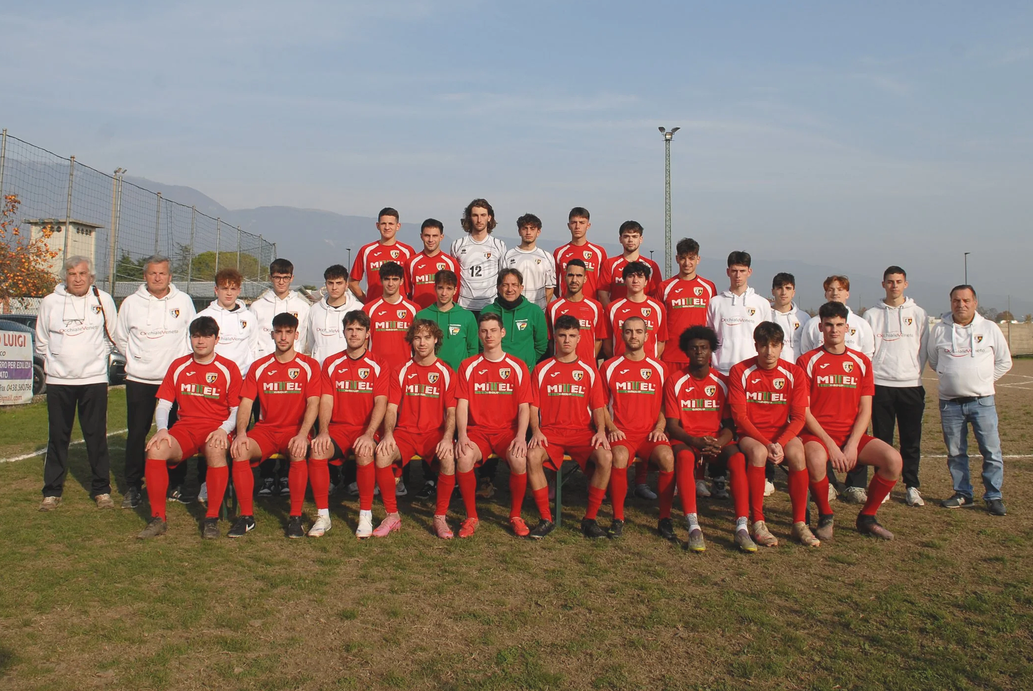 Gruppo di calcio con giocatori in divisa rossa e bianca, allenatori e staff fotografati sul campo di calcio.